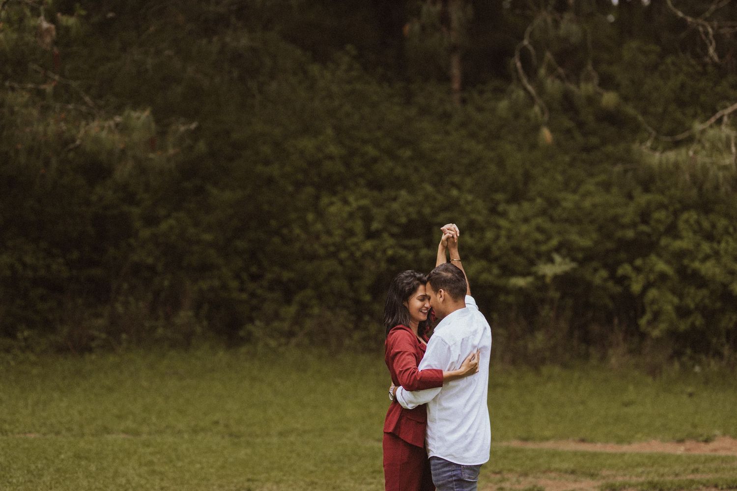 A couple in maroon and white outfits dance together in an outdoor setting surrounded by greenery.