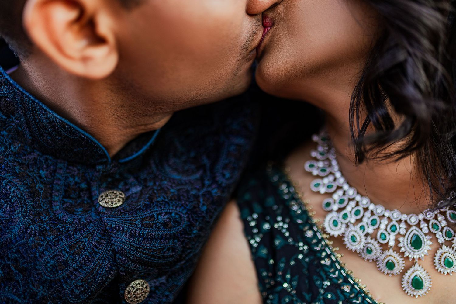 A romantic couple in navy blue outfits sharing an intimate kiss with a sparkling necklace visible.