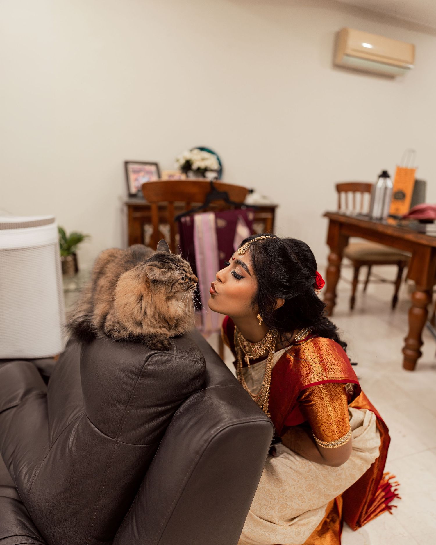 Woman in red and gold saree sharing a sweet moment with a fluffy cat on a leather sofa indoors.