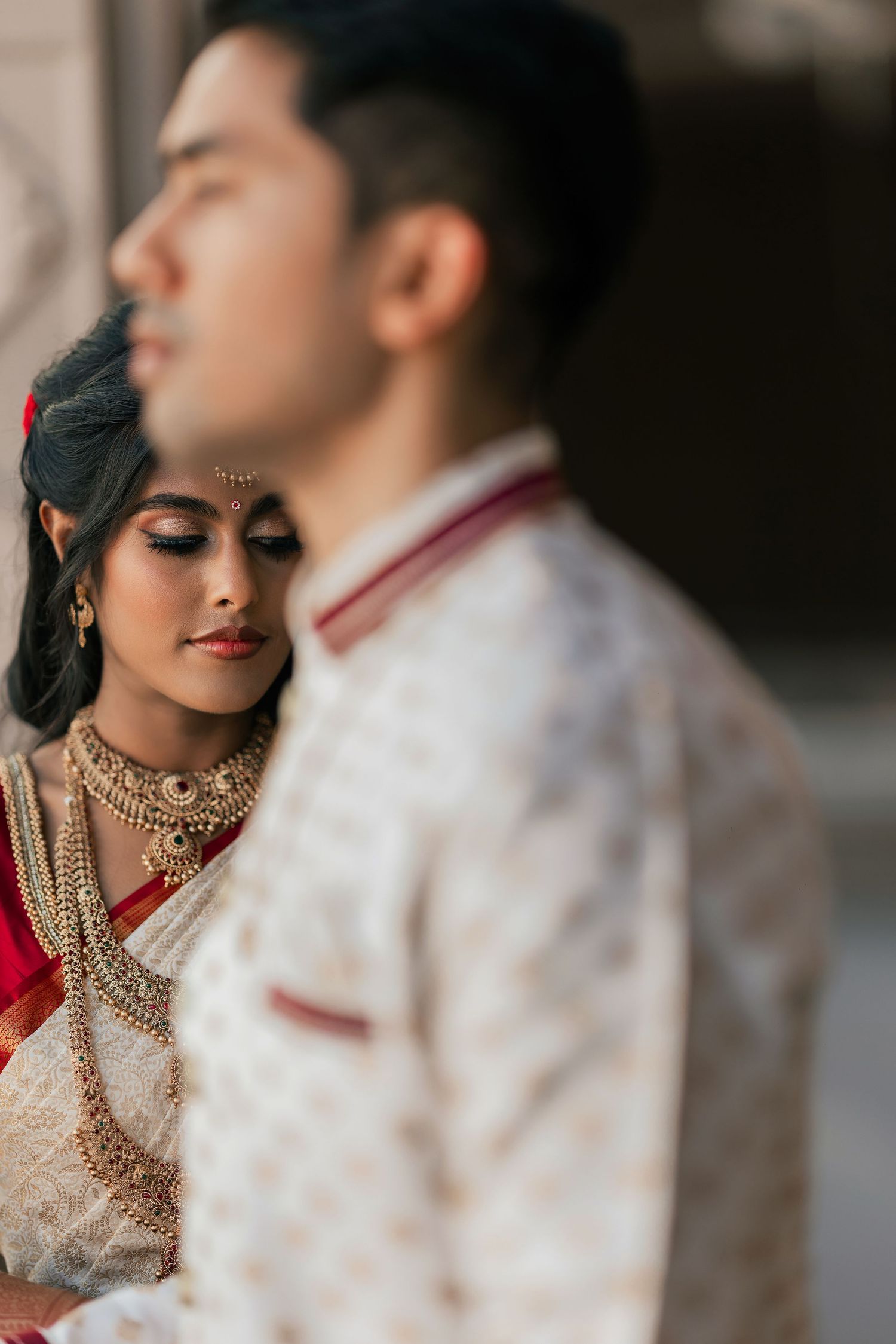 Indian bride in red saree with gold jewelry leans on groom in white sherwani during traditional wedding ceremony.