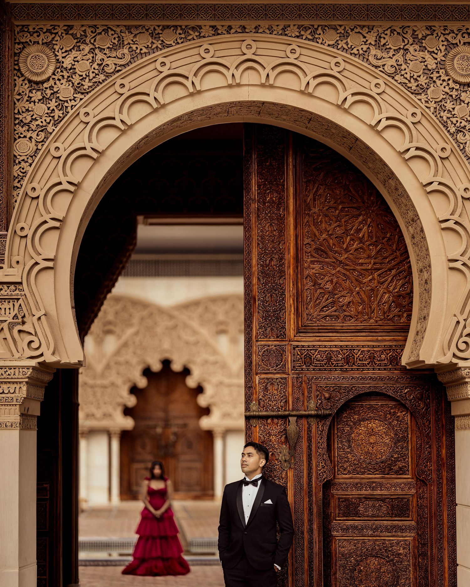 Man in tuxedo stands by ornate Moroccan arch door, woman in red gown posed in background, elegant pre-wedding shoot.