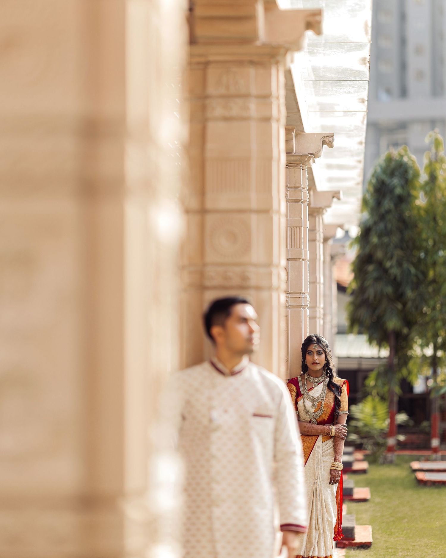 Indian bride in white and gold saree stands between ornate columns as groom in white sherwani poses in foreground.