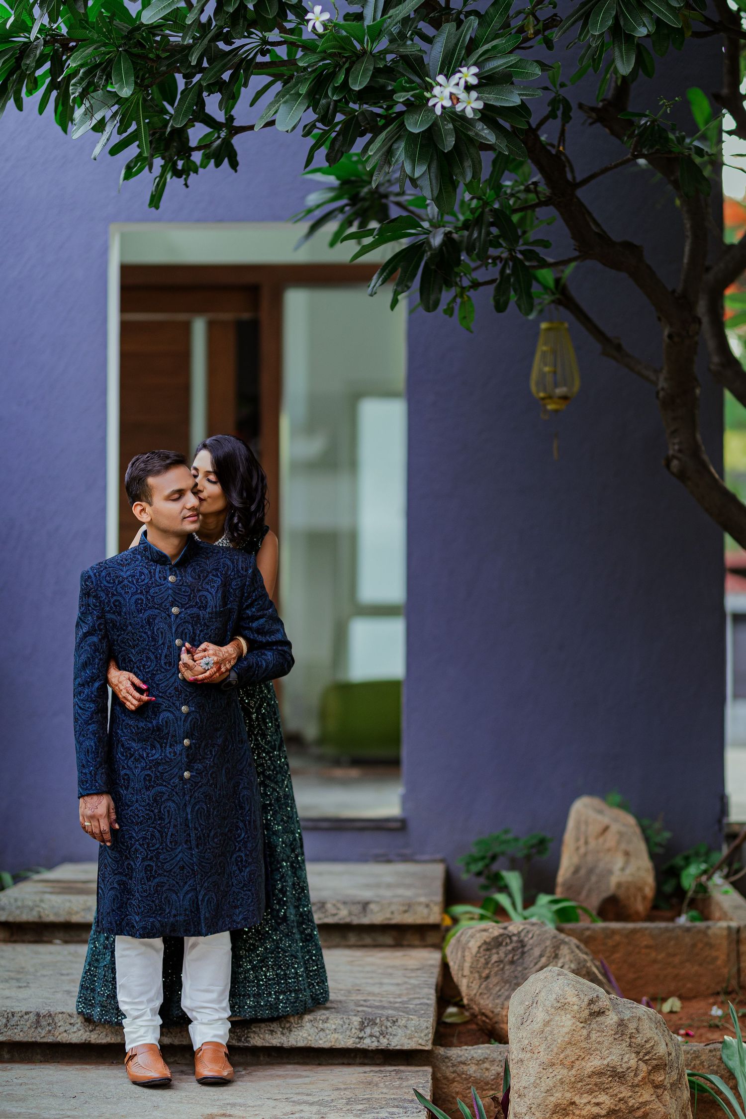 A couple in coordinating navy blue outfits share an intimate moment on stone steps surrounded by greenery.