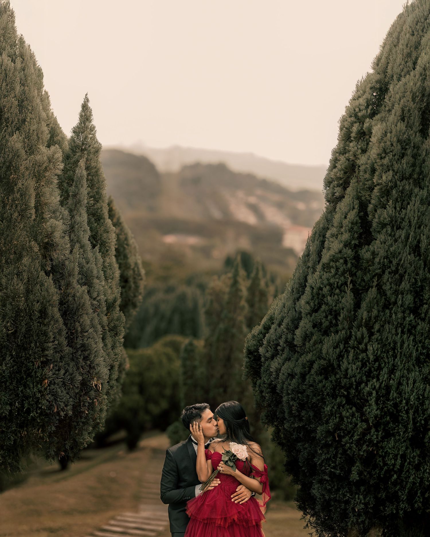Couple kissing among tall cypress trees with misty hills, woman in red dress, romantic outdoor engagement photo.