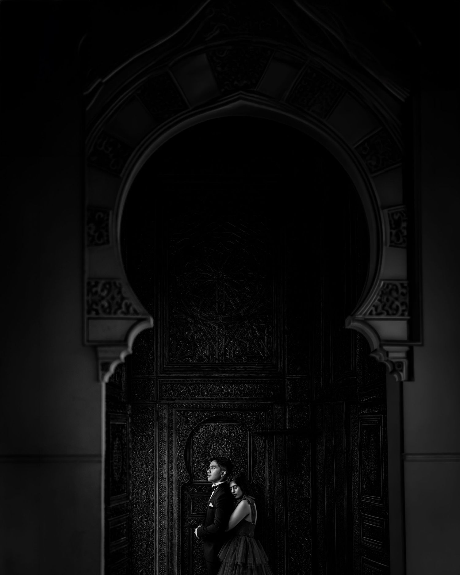 Black and white photo of a couple embracing under an ornate Moorish arch with intricate carved details.