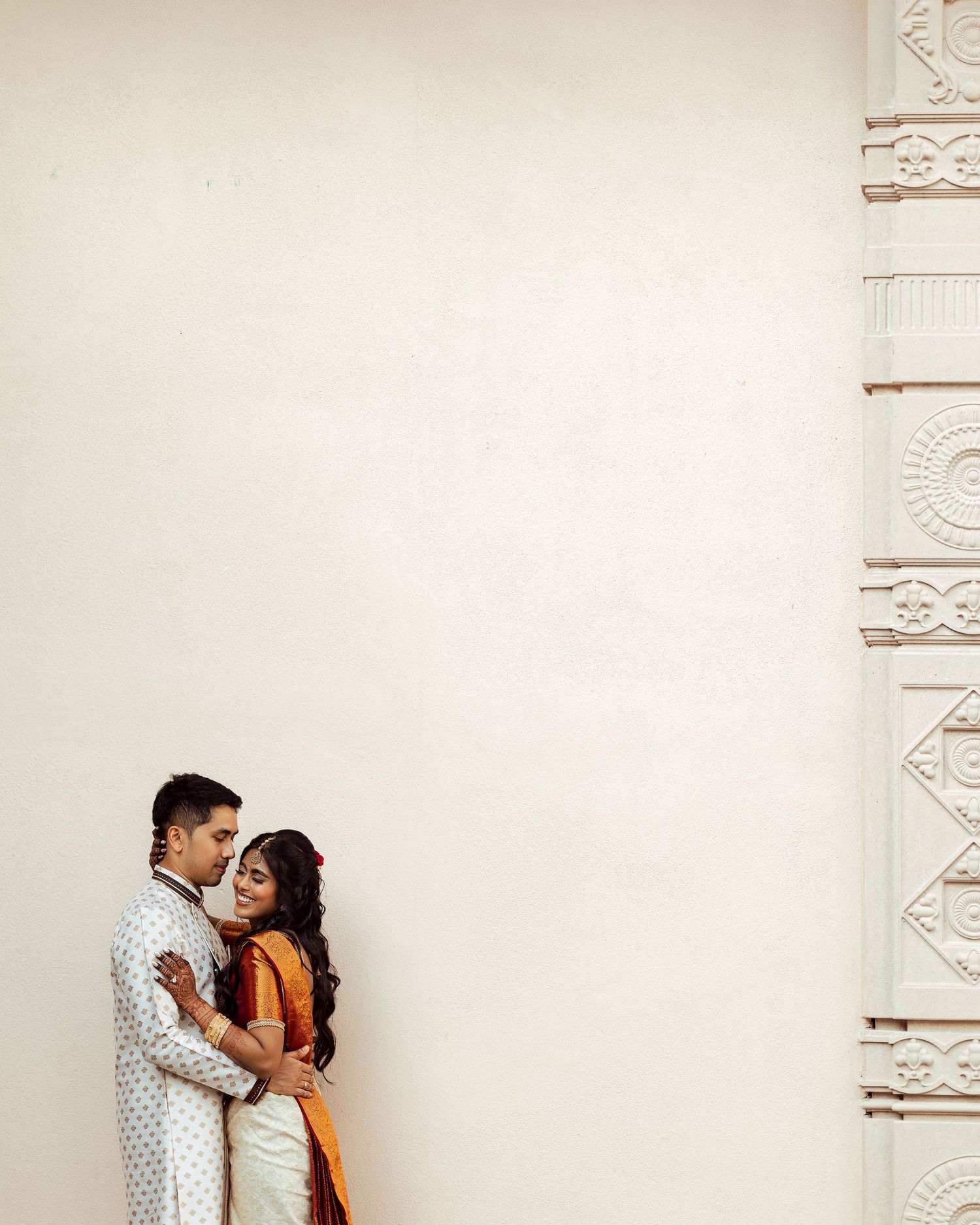 A couple in traditional Indian attire embraces tenderly against a white wall with ornate architectural details.