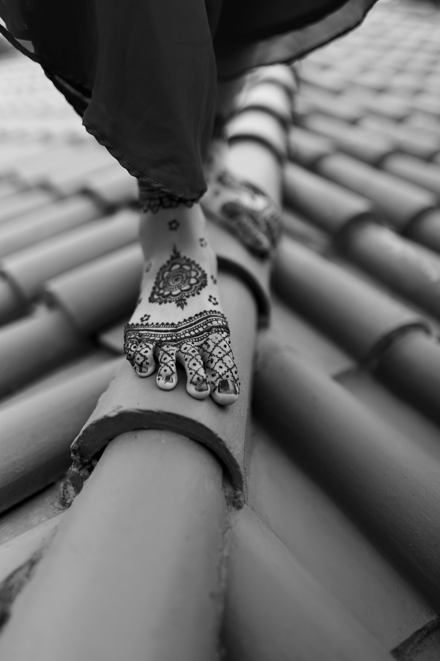 Black and white photo of a foot with intricate henna design walking on curved terracotta roof tiles.