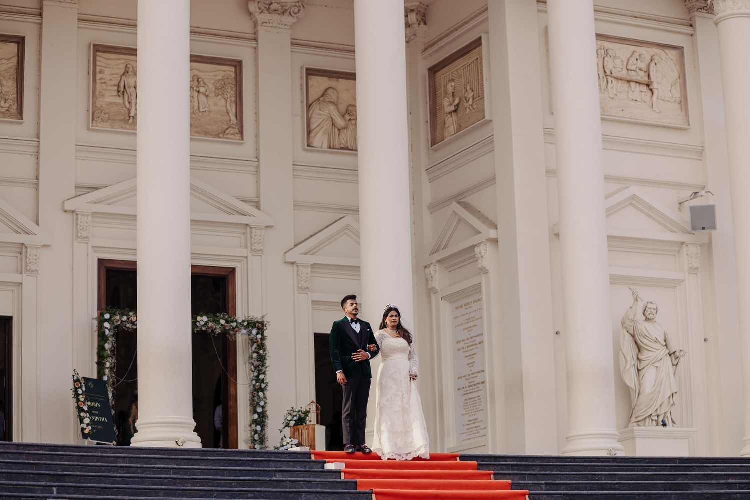 A couple stands on ornate white marble steps with classical columns during a wedding ceremony at a grand venue.