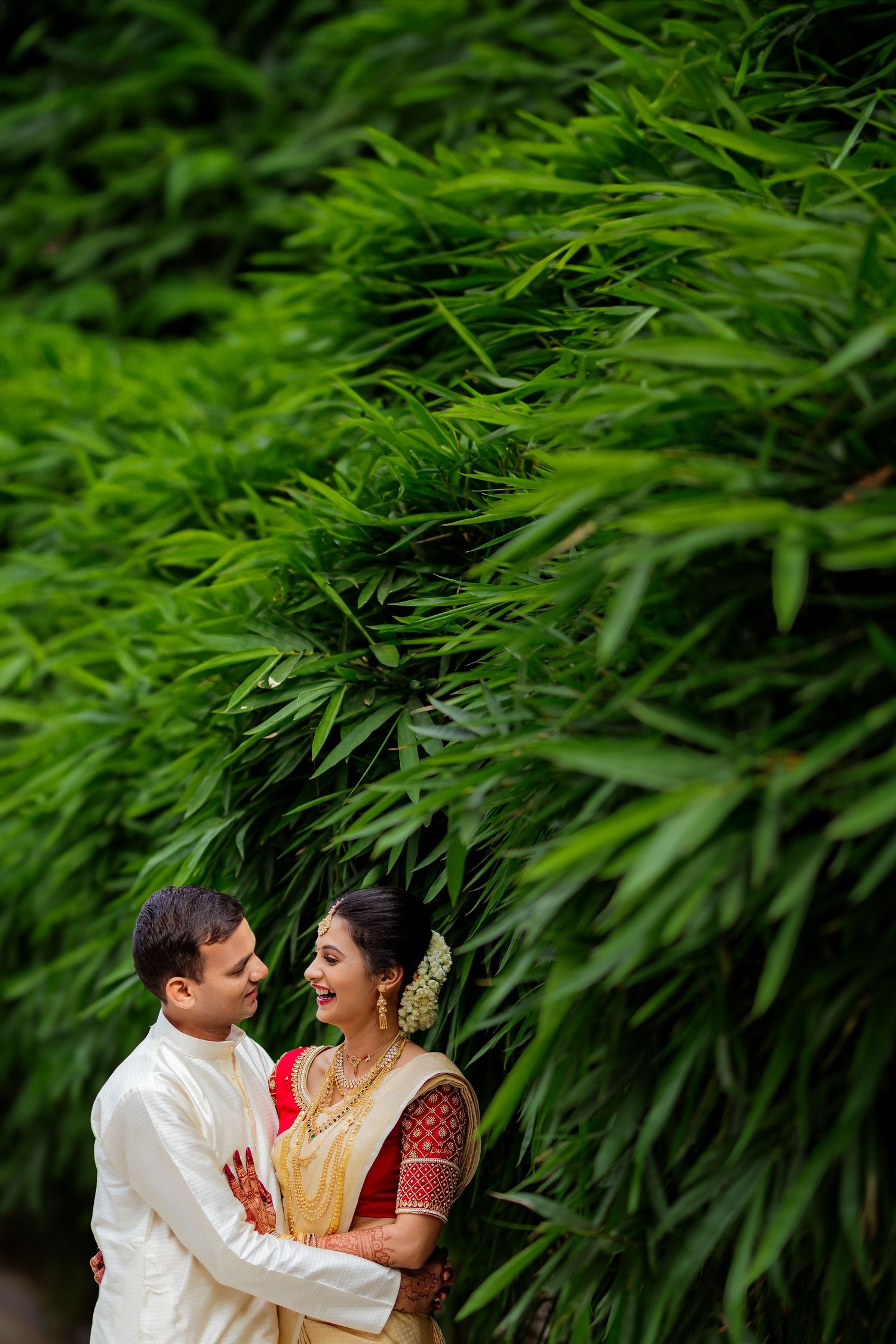 Couple in traditional Indian attire poses together against lush green bamboo foliage background.