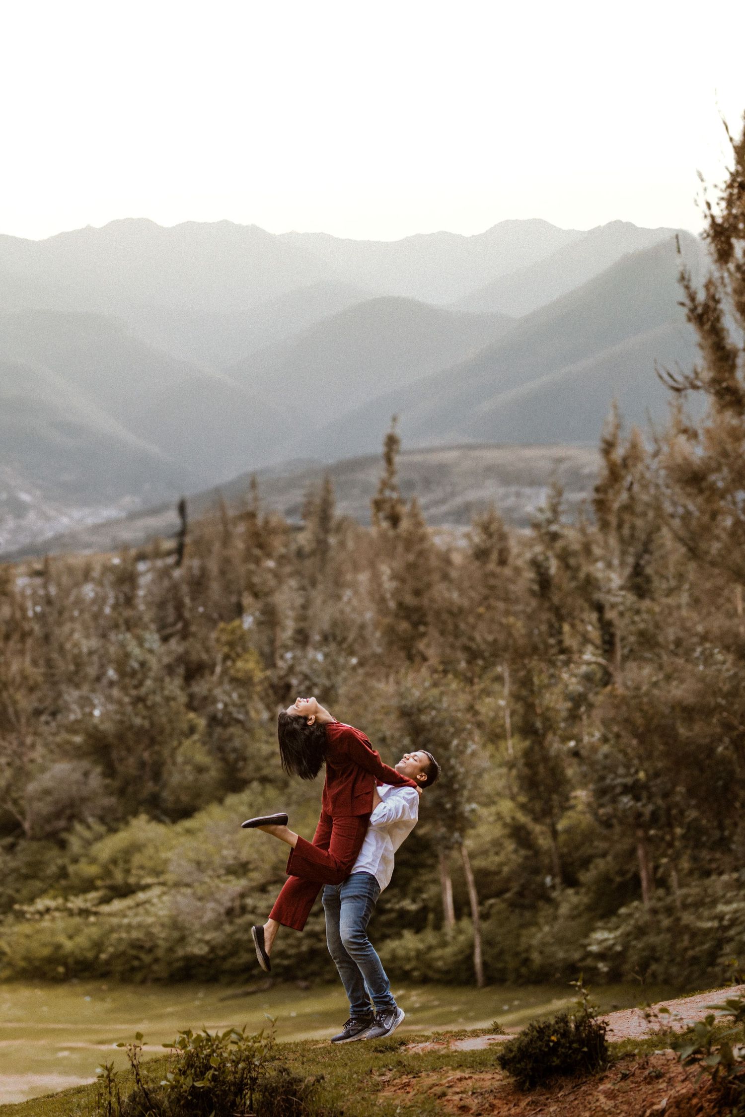 A person in a red jacket bends over to take a photograph on a scenic mountain trail with misty peaks in the background.