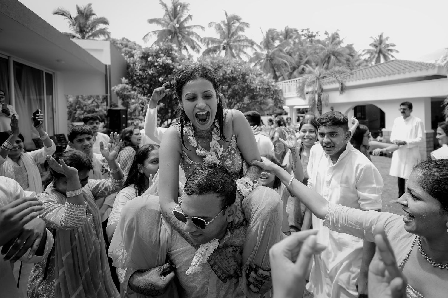 People dance and celebrate together at an outdoor party with palm trees in the background.