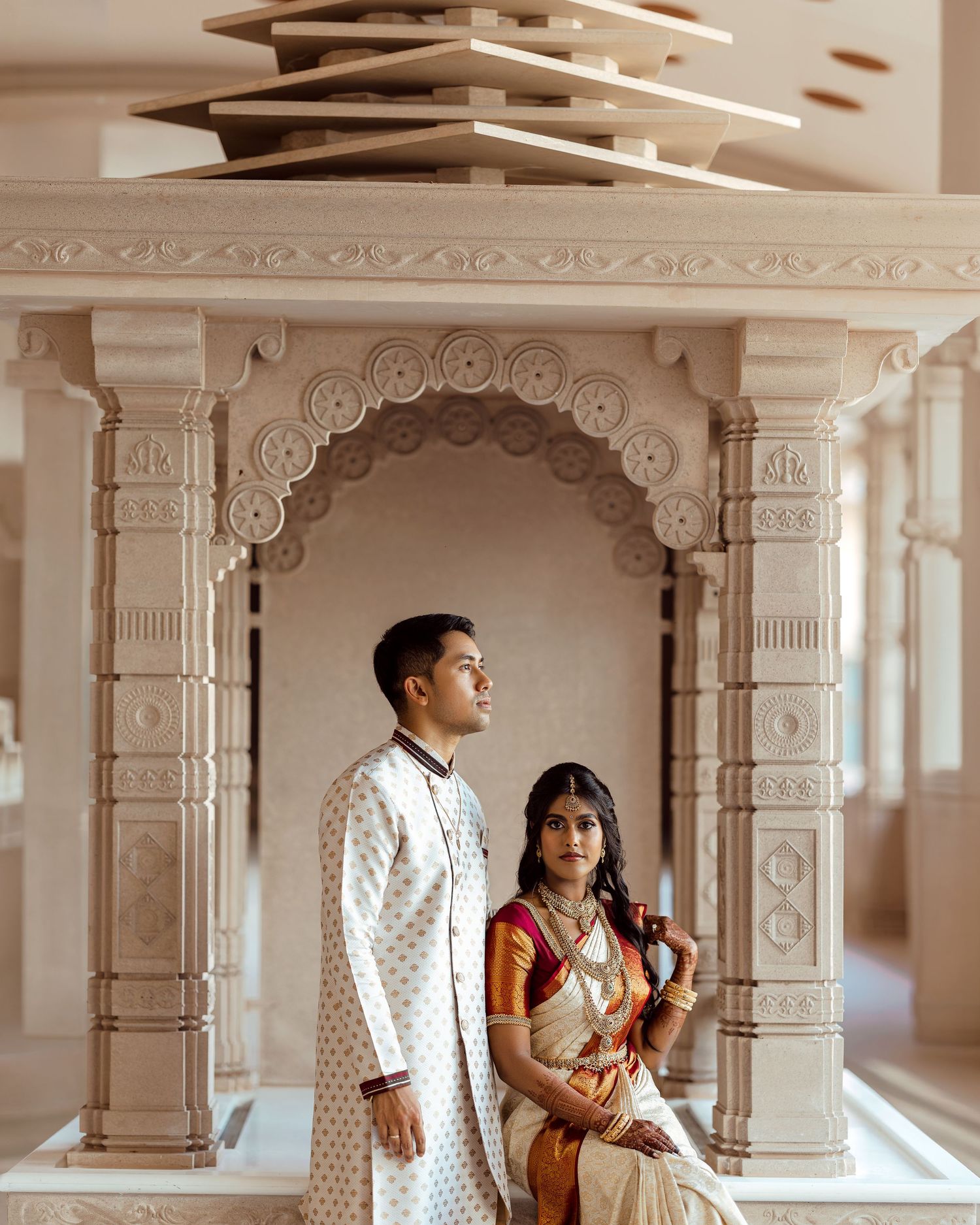 Indian couple in traditional wedding attire posing elegantly in an ornate temple archway with intricate stone carvings.