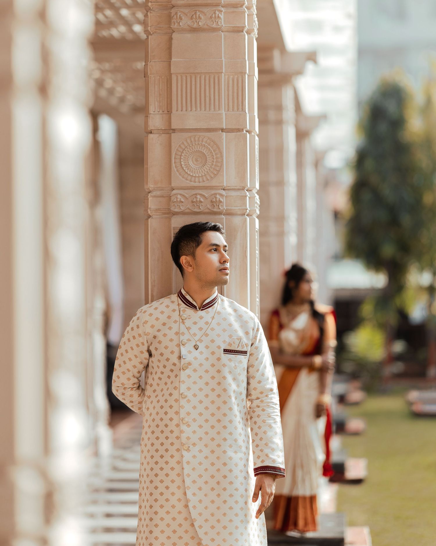 A man in a white embroidered kurta stands by ornate columns, with a woman in a saree visible in the background.