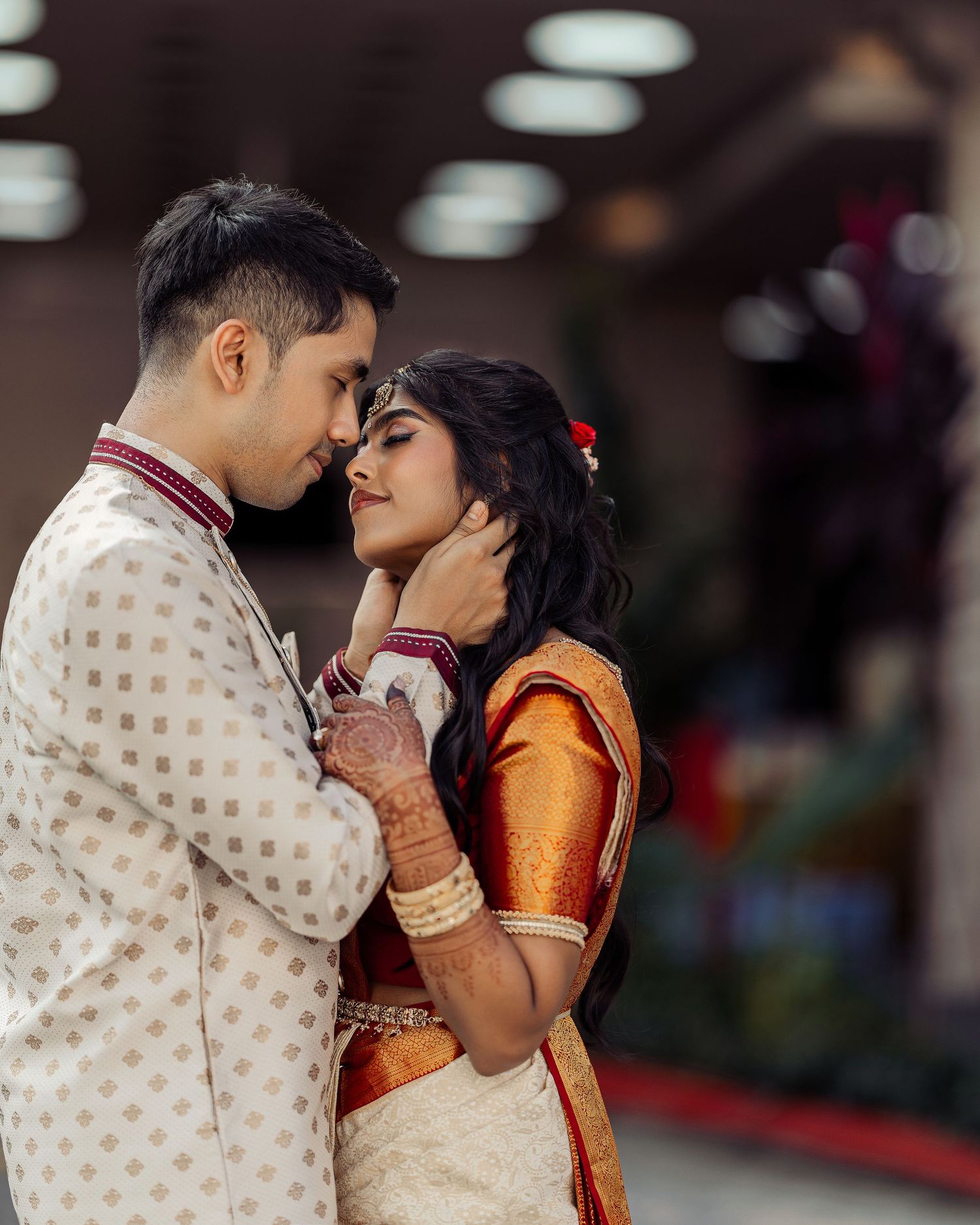 Indian couple in traditional wedding attire sharing a romantic moment, bride in orange saree with gold jewelry.