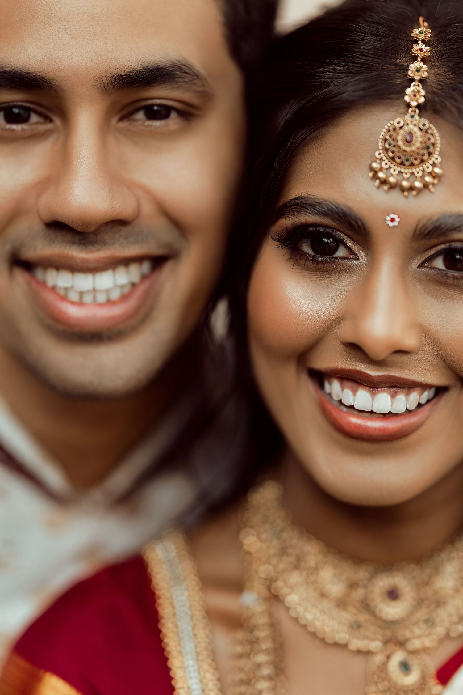 Smiling Indian bride and groom in traditional wedding attire, bride wearing gold maang tikka and necklace.