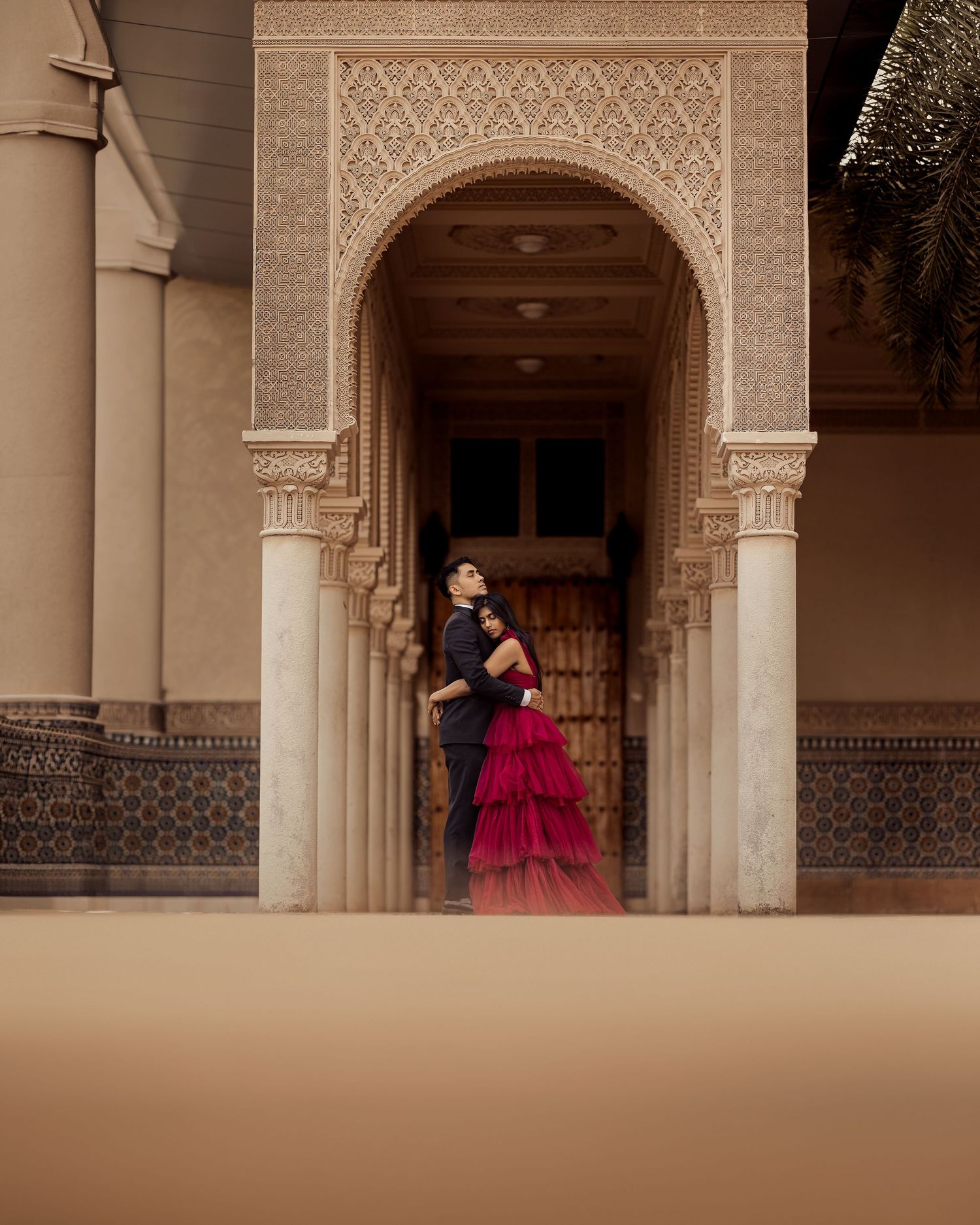 Couple embracing in elegant Moorish archway, woman in red tiered gown, ornate carved architecture backdrop.