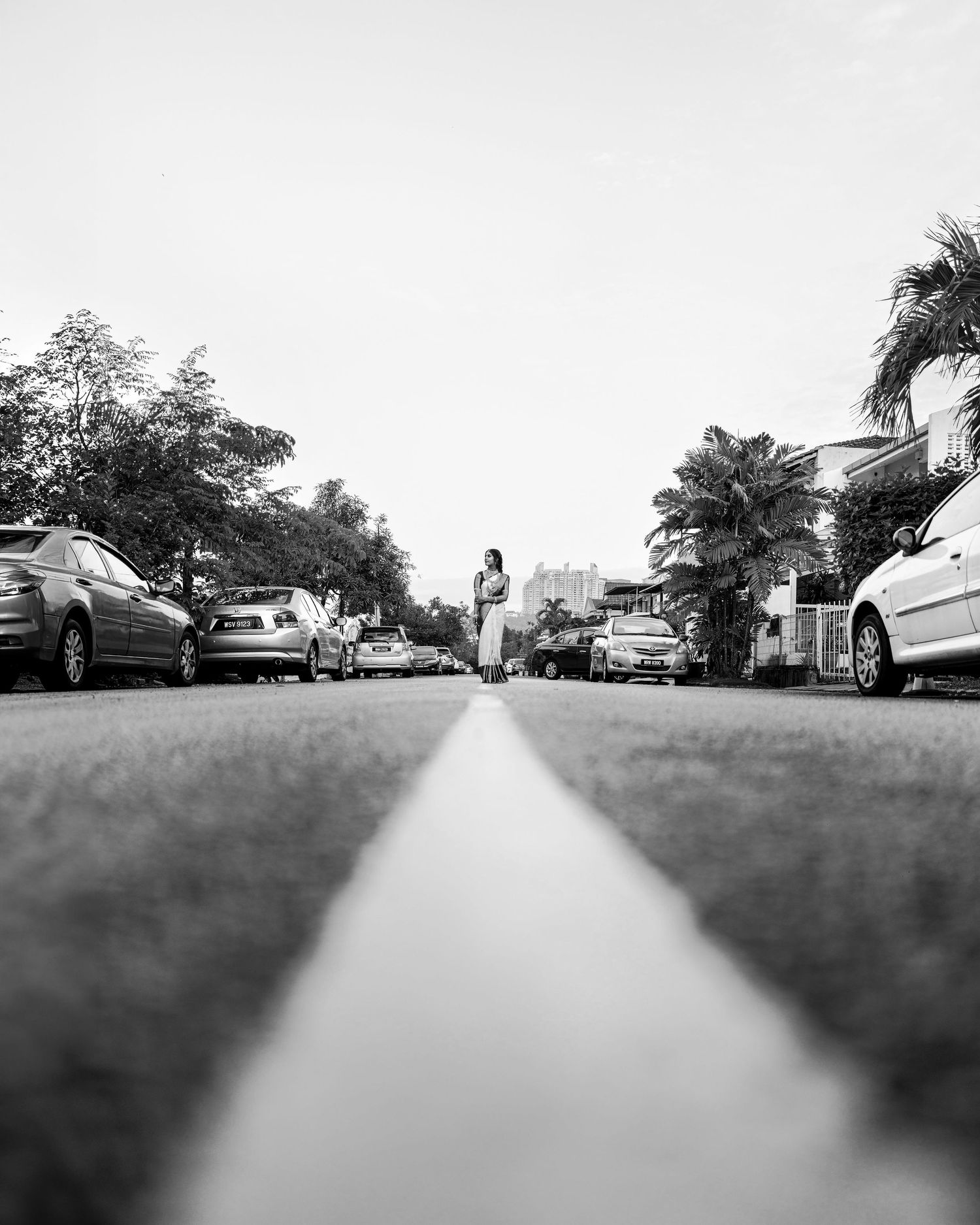 Black and white low-angle street photo with parked cars on both sides and a person standing in the middle of the road.