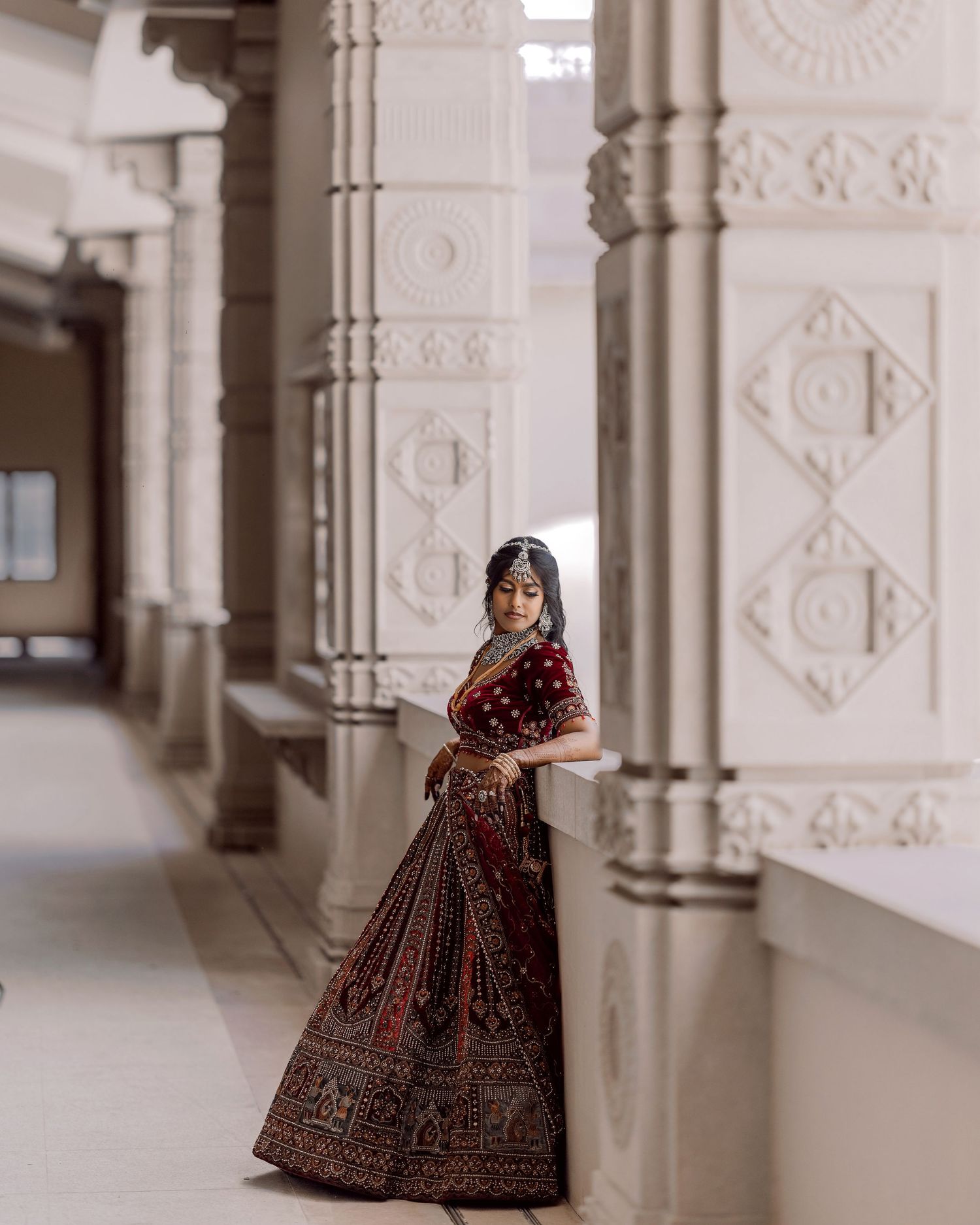 A woman in a stunning dark red embroidered lehenga poses gracefully between ornate white pillars of an elegant building.