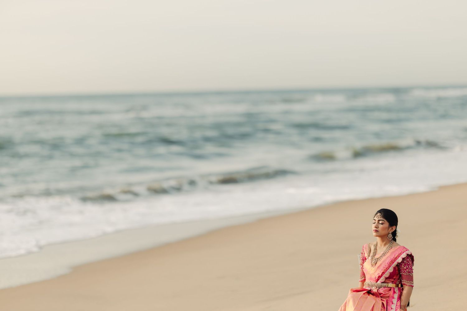 Couple walking along sandy beach shore in traditional Indian wedding attire during golden hour.