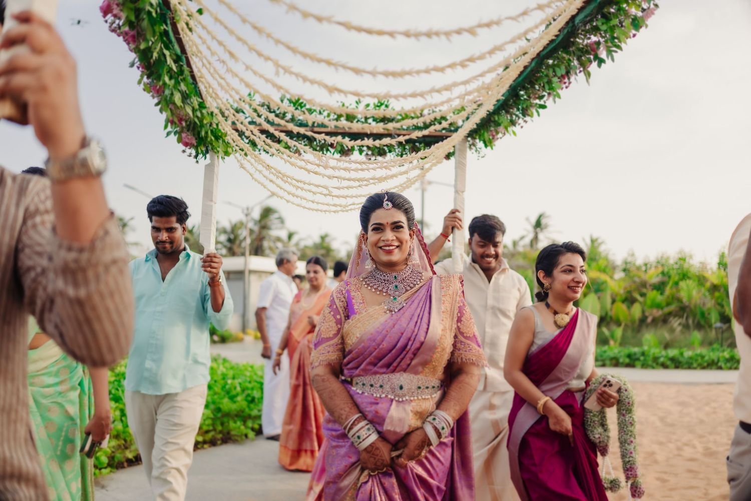 Guests gather under elegant white draped canopies with greenery decorations at an outdoor wedding ceremony.