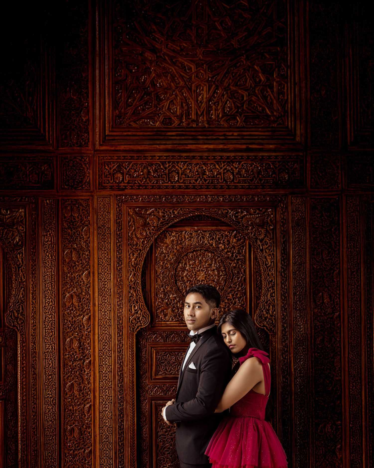A romantic couple poses against an ornate wooden carved wall backdrop in formal evening attire.
