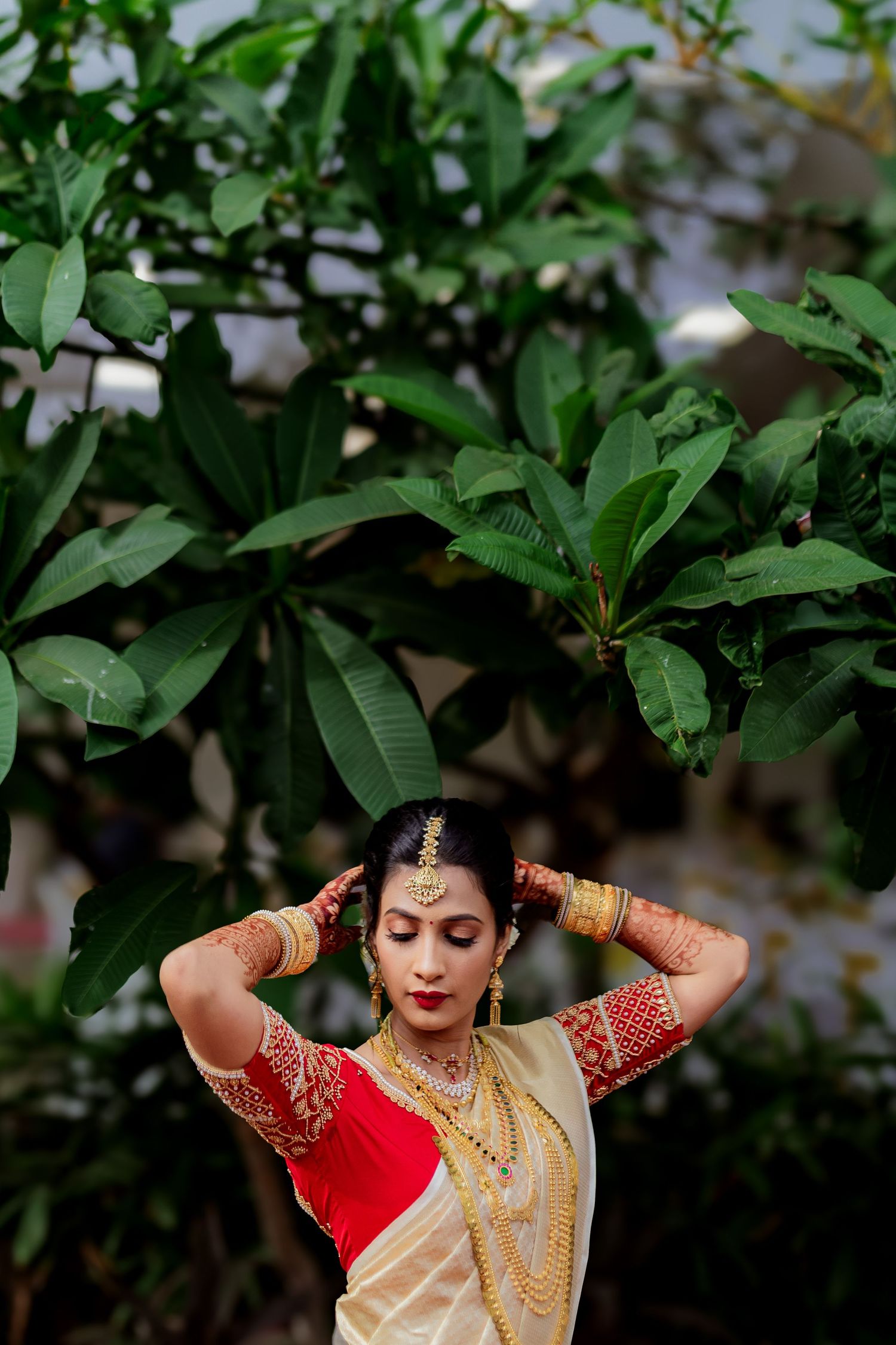 A person in traditional South Asian bridal attire with gold jewelry poses against green foliage background.