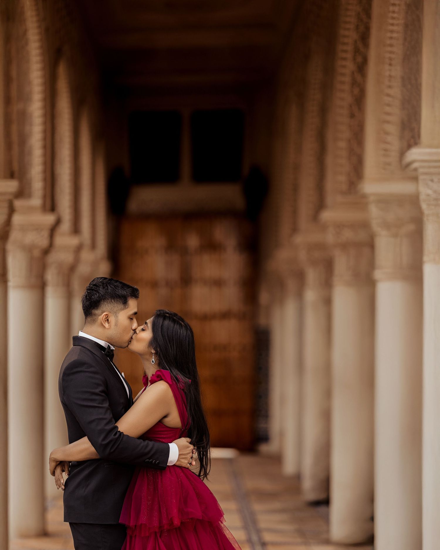 Couple in formal attire embracing during romantic photoshoot at ornate stone archway colonnade.