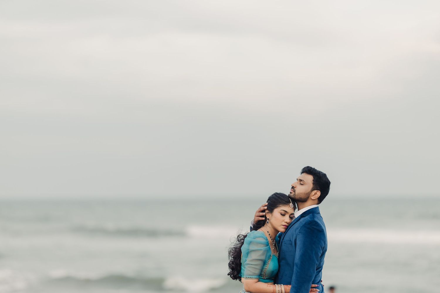 A couple embraces on a misty beach with gray skies and ocean waves in the background.
