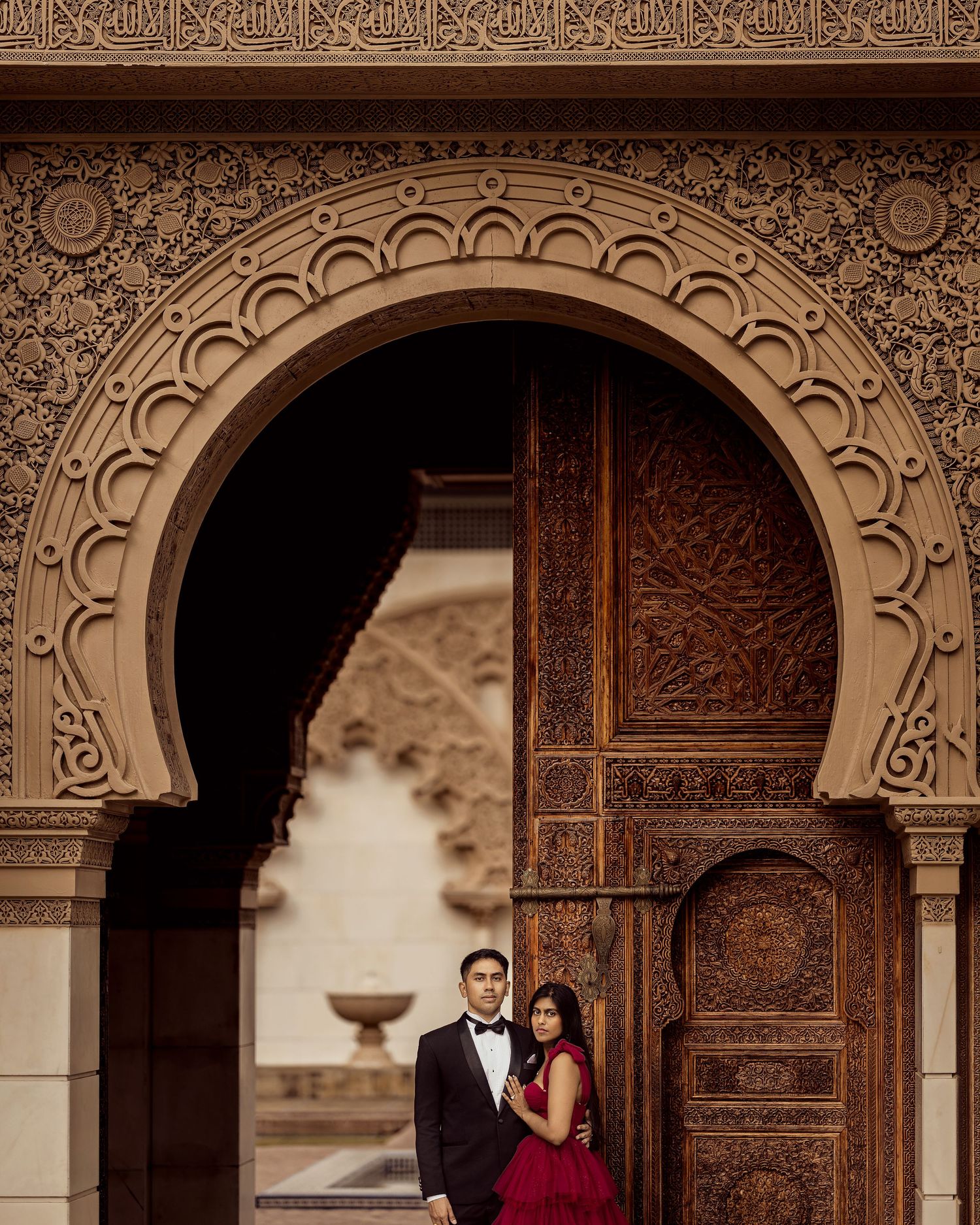 Couple in formal attire poses under ornate Moroccan arch with intricate carved wooden doors.