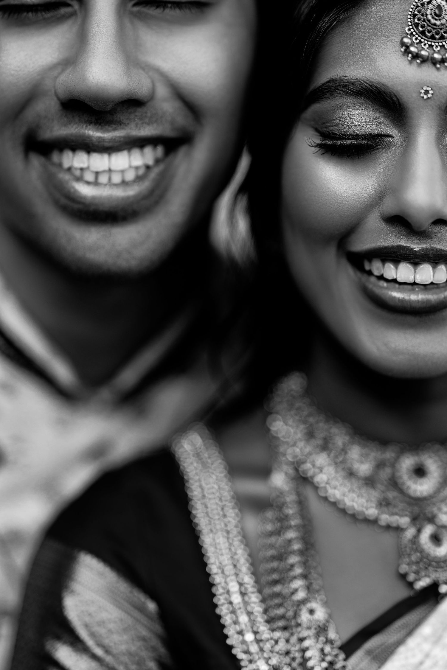 Black and white portrait of smiling Indian bride and groom in traditional wedding attire and jewelry.