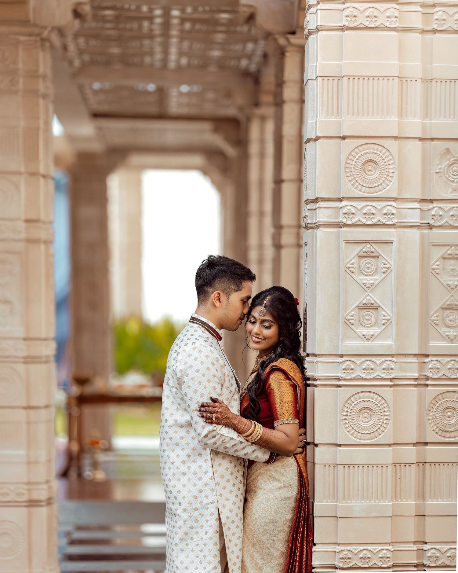 Couple in traditional Indian wedding attire embracing by ornate white temple pillars.