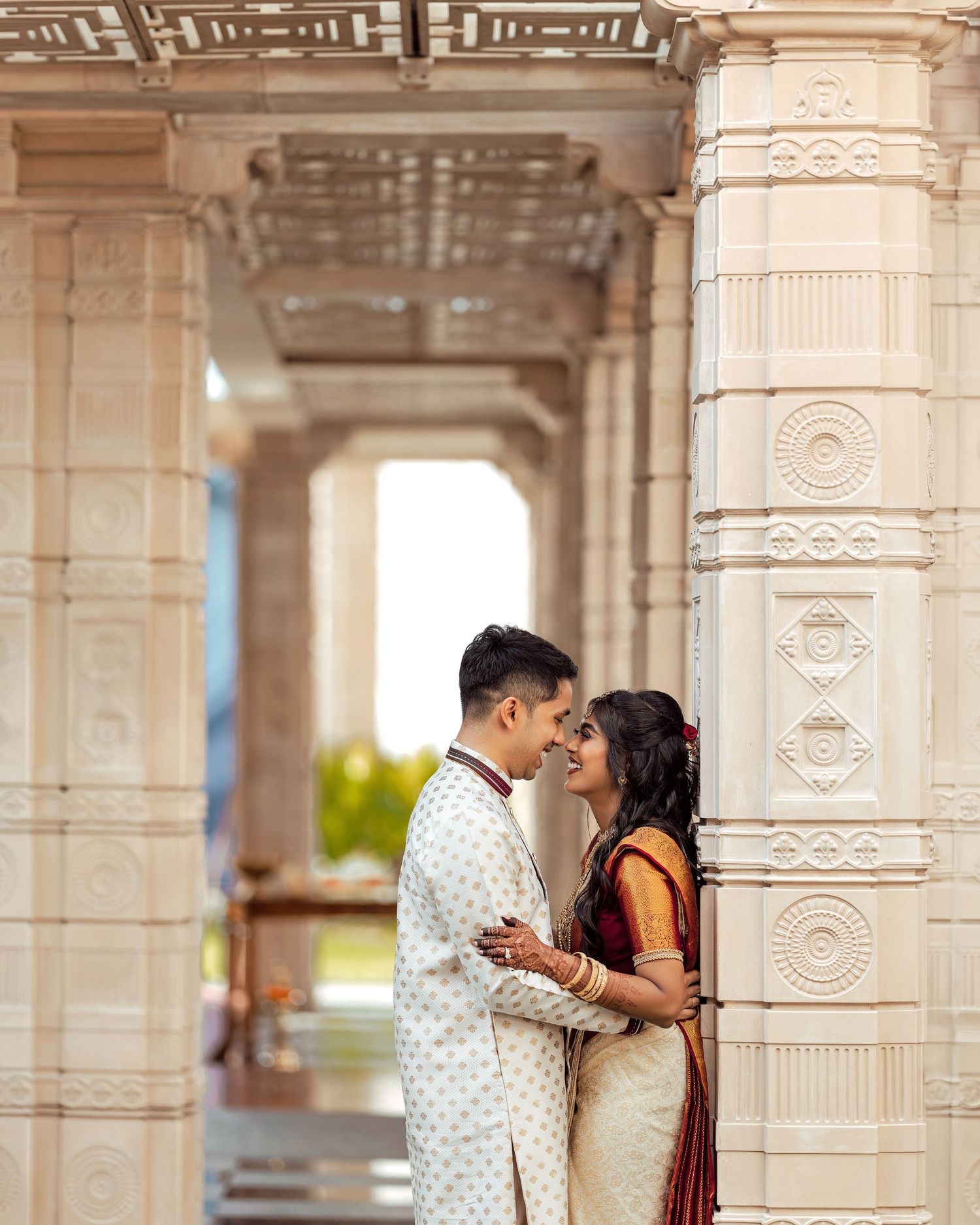 Indian couple in traditional wedding attire sharing a romantic moment between ornate white marble temple columns.