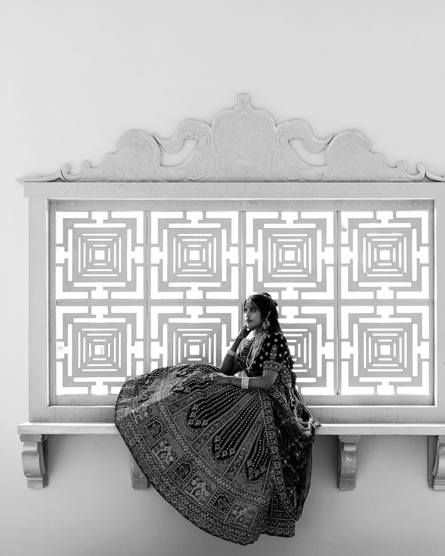 Black and white photo of a woman in an ornate lehenga sitting before a decorative geometric lattice panel.