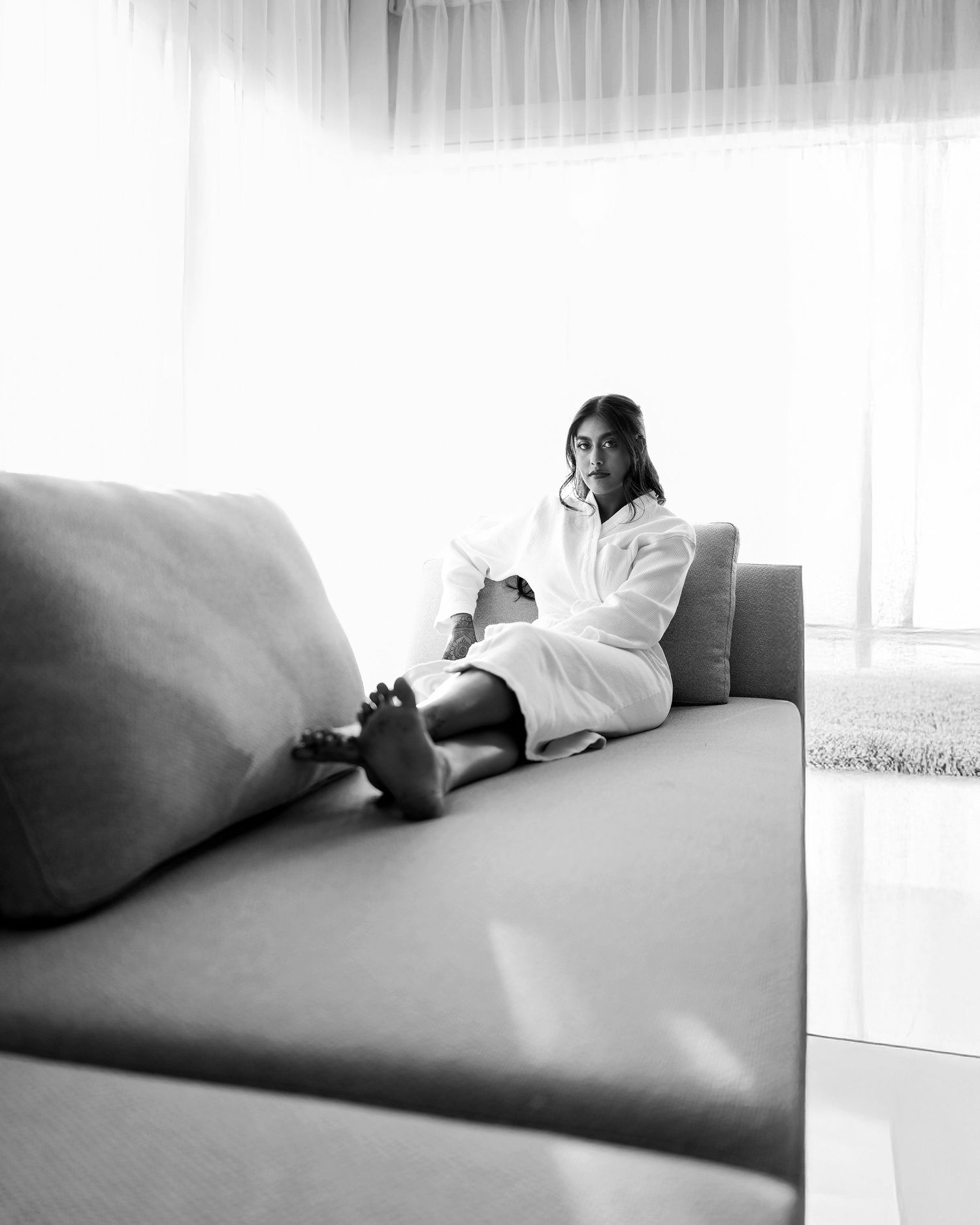 Black and white photo of a woman in a white robe sitting relaxed on a modern sofa near sheer curtains.