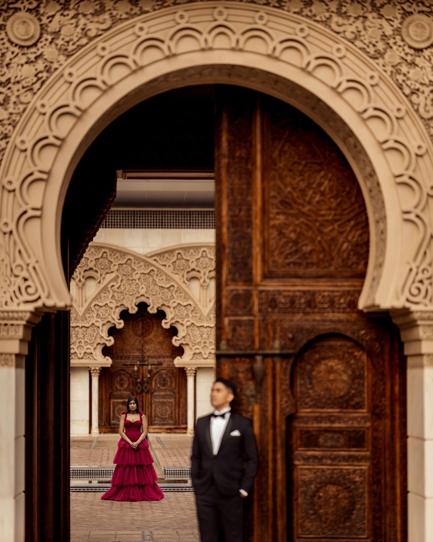 Couple posing in elegant Moroccan archway with intricate carved details, woman in red gown, man in tuxedo.
