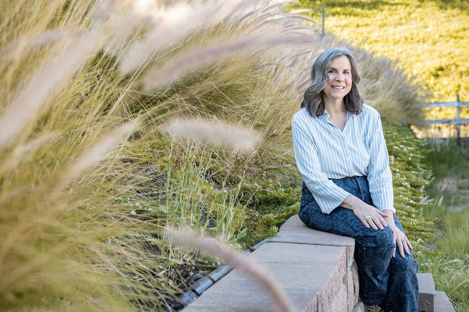 Woman with gray hair sits on stone wall surrounded by ornamental grasses in a sunlit garden setting.