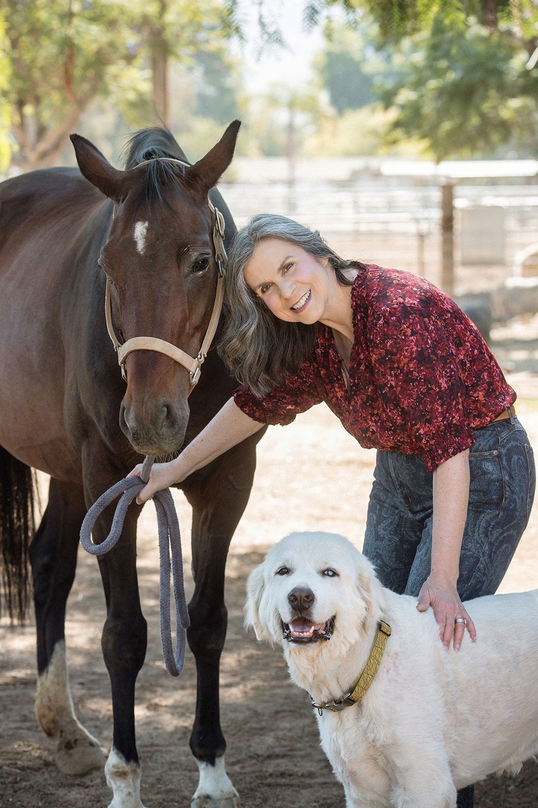 Smiling woman poses with a brown horse and white fluffy dog at an outdoor ranch setting.