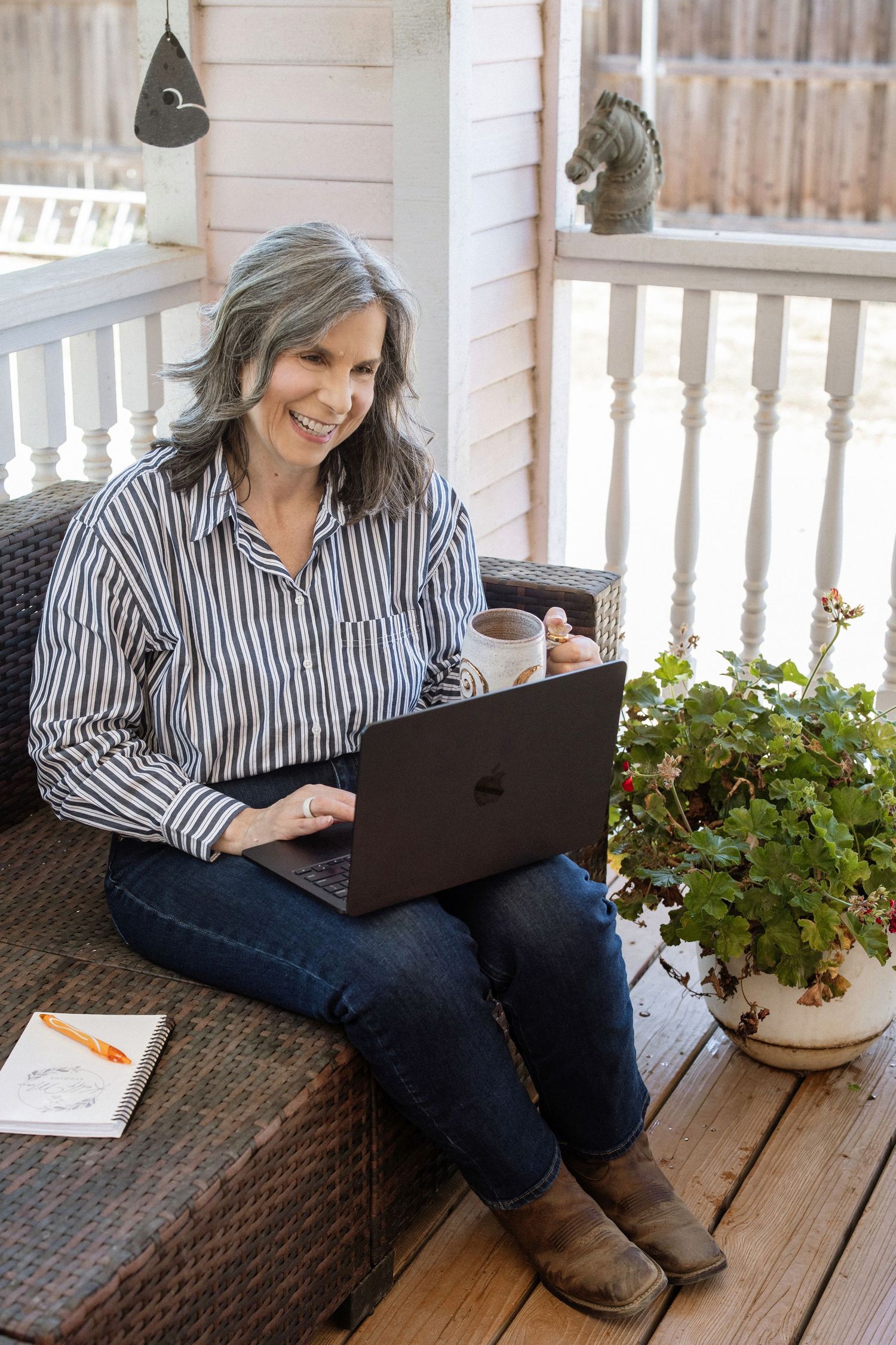 Smiling middle-aged woman with gray hair working on laptop on porch, holding coffee mug, wearing striped shirt and jeans.