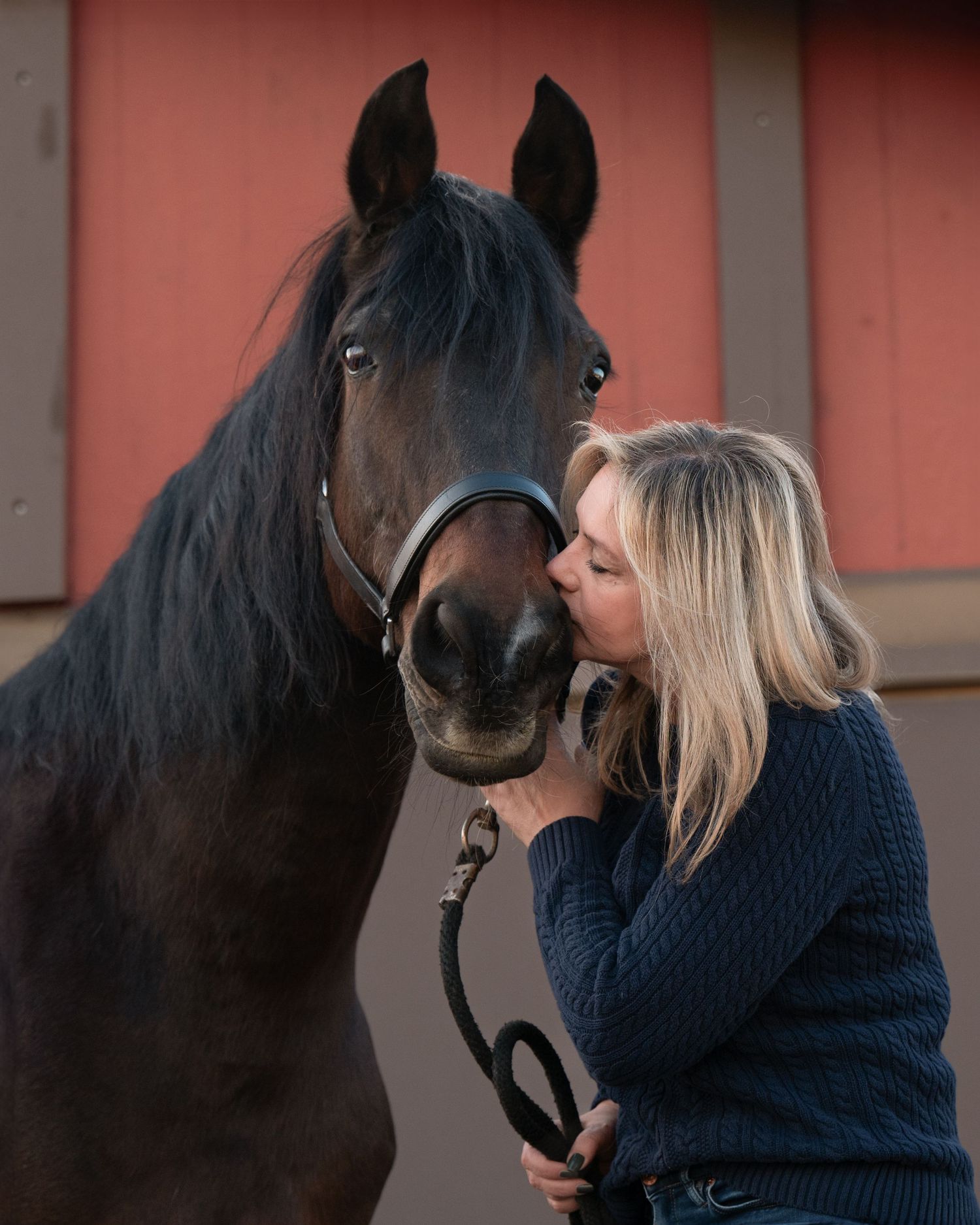 A blonde woman in a navy sweater shares a tender kiss with a dark bay horse near a red barn.