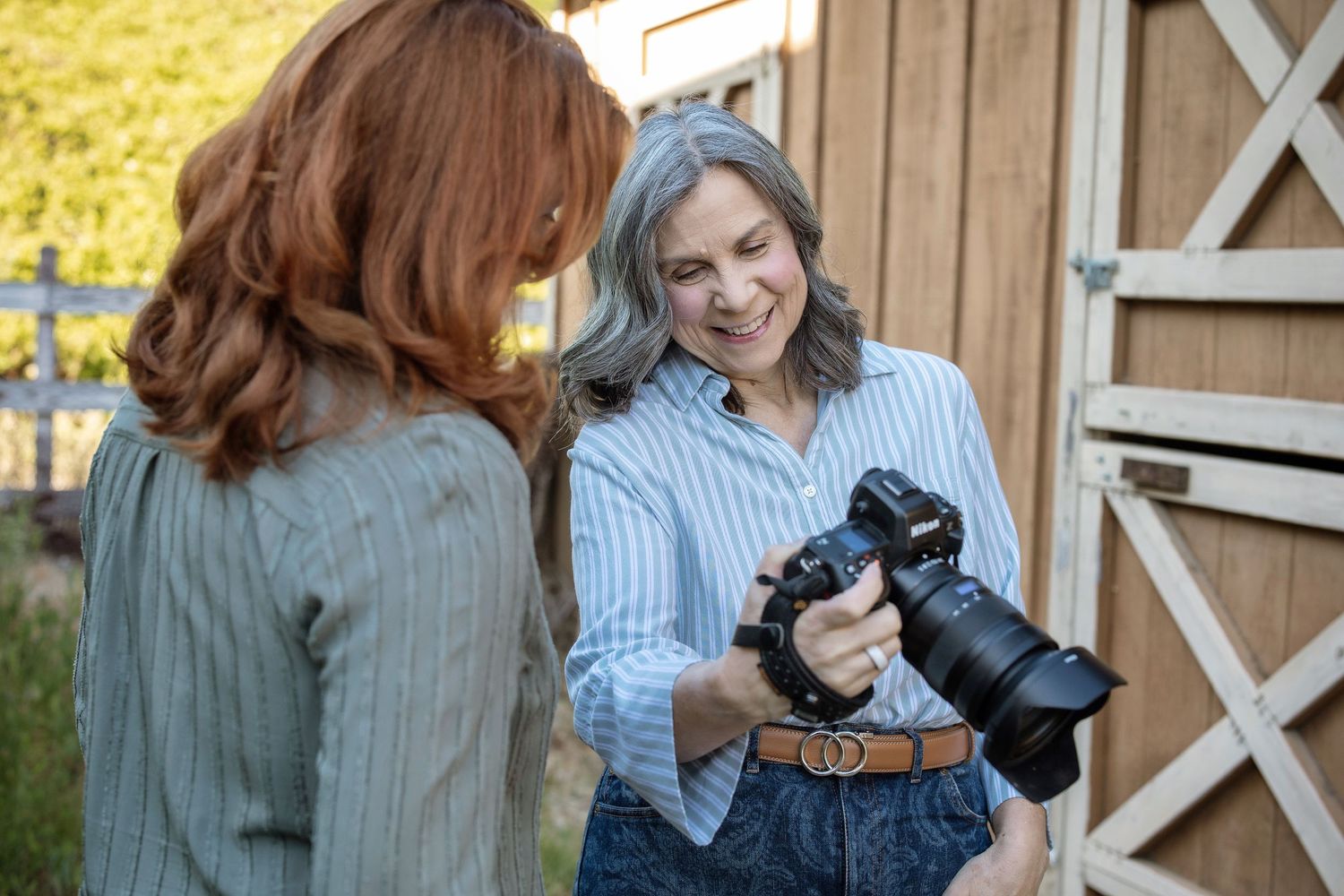 Two women examine photos on a camera outside a rustic barn, one holding a DSLR camera while smiling.
