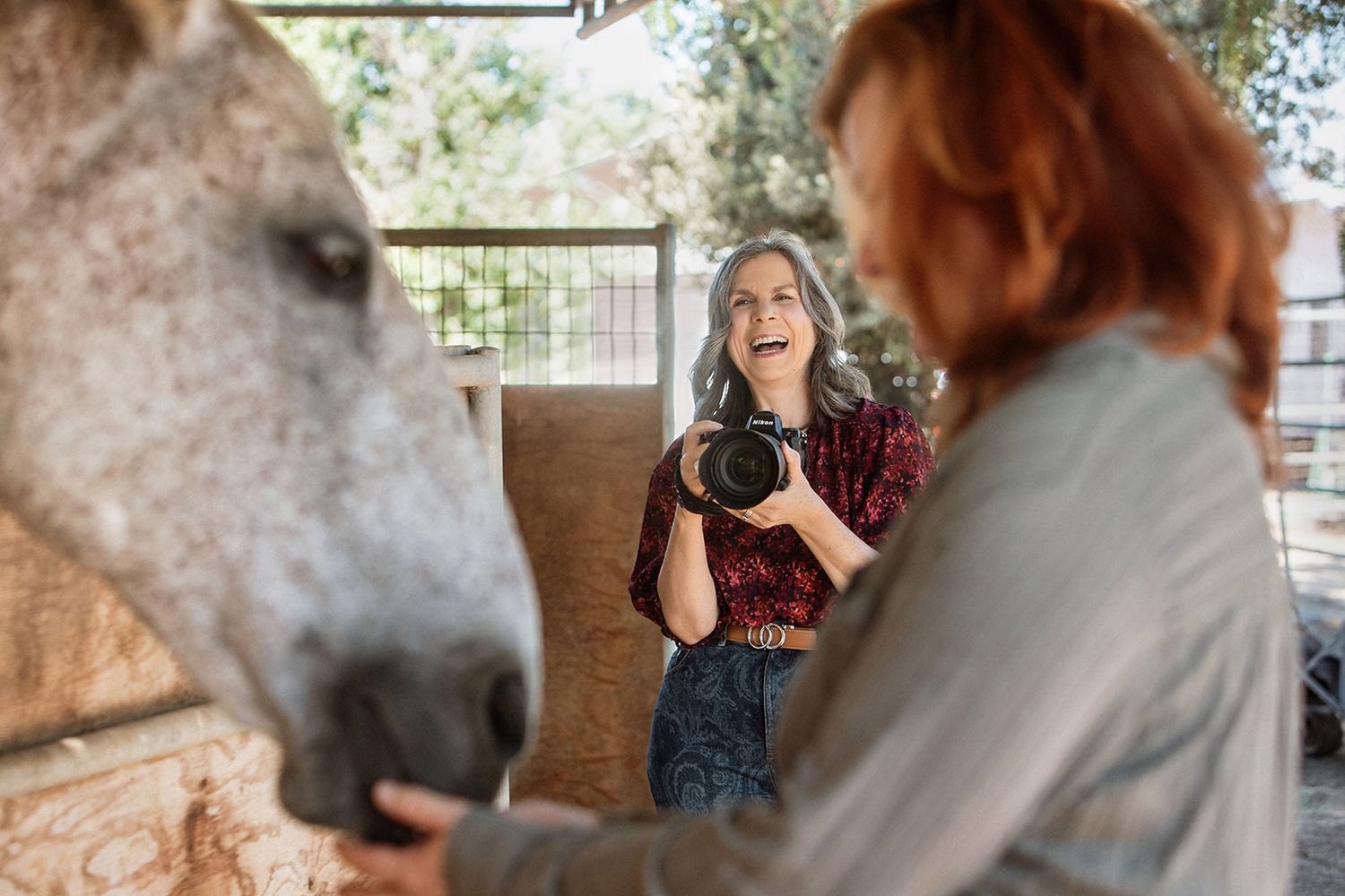 Photographer laughing while capturing woman petting a gray horse in a rustic stable setting.