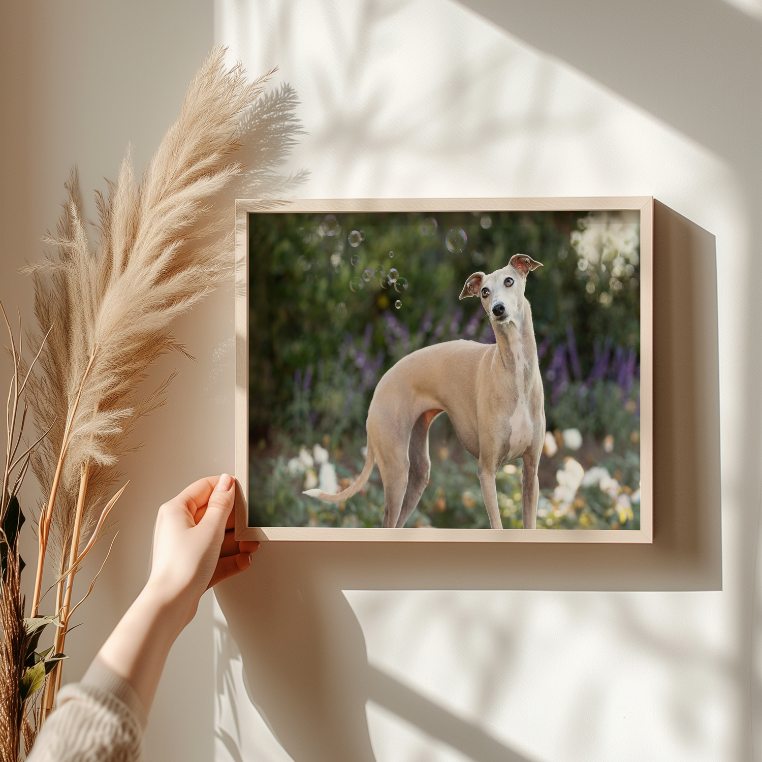 Hands arranging dried pampas grass stems near framed artwork in sunlit room.