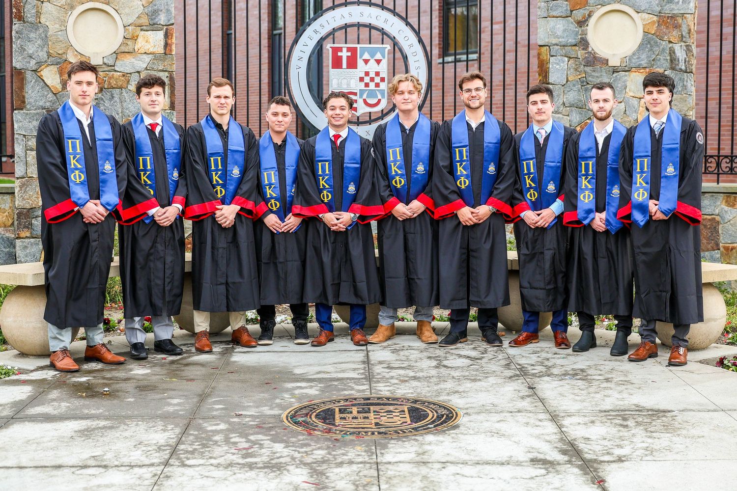 A line of graduates in black robes and colorful stoles pose for a group photo in front of a brick wall.