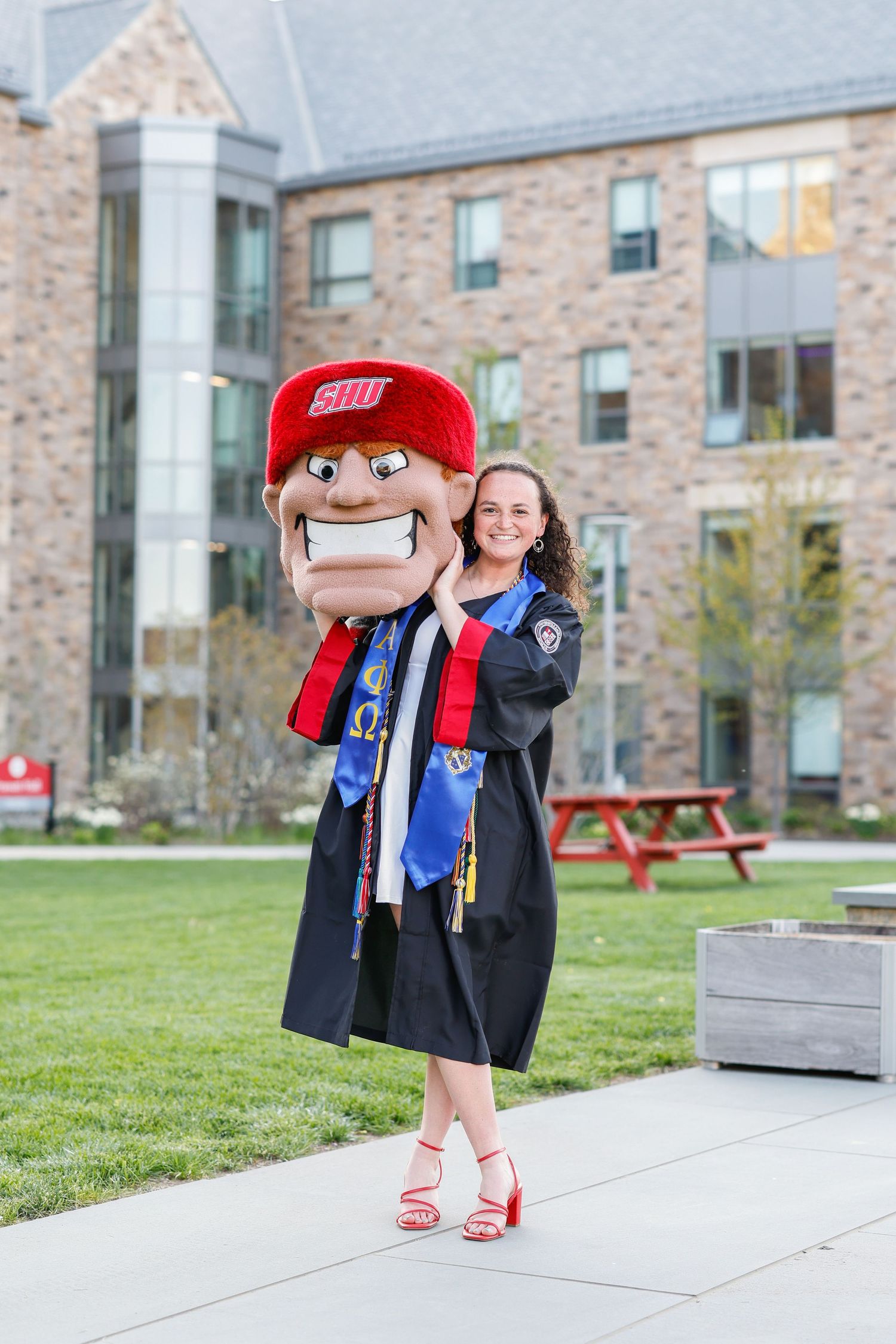 A graduate poses with a university mascot plush toy on campus grounds.