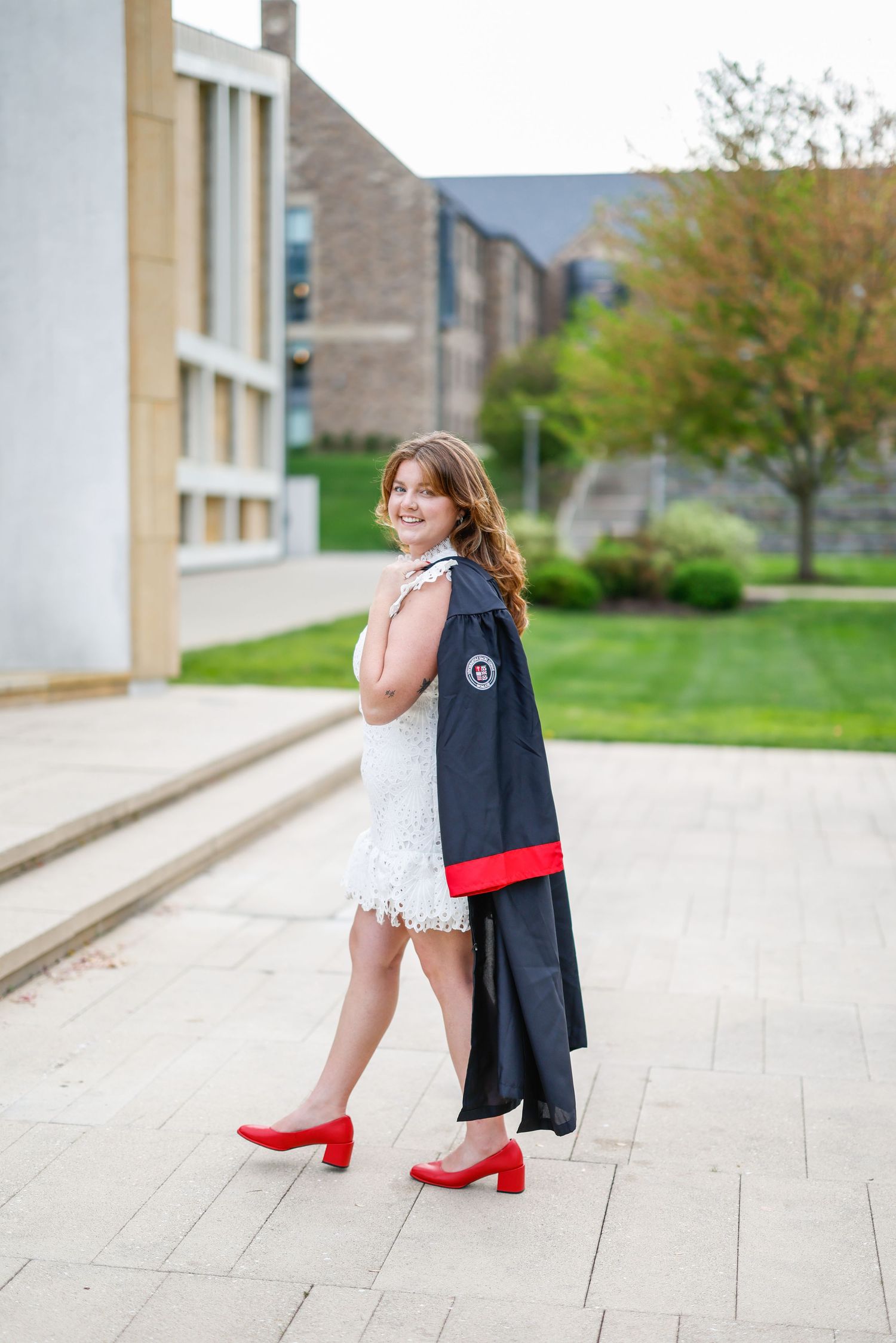 Graduate in navy blazer and red heels poses on campus sidewalk near academic buildings.