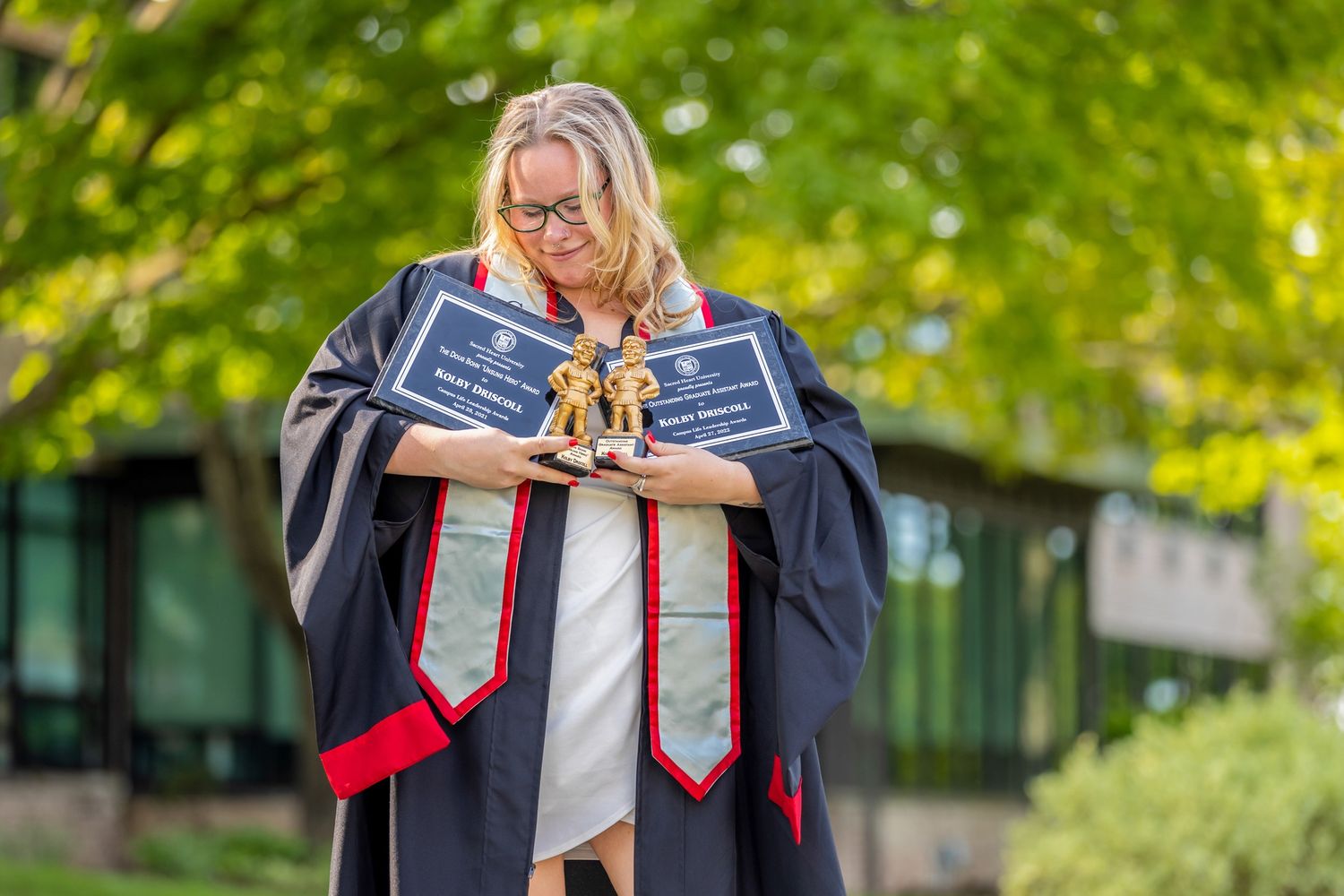 Graduate in academic regalia posing outdoors with trees in background.