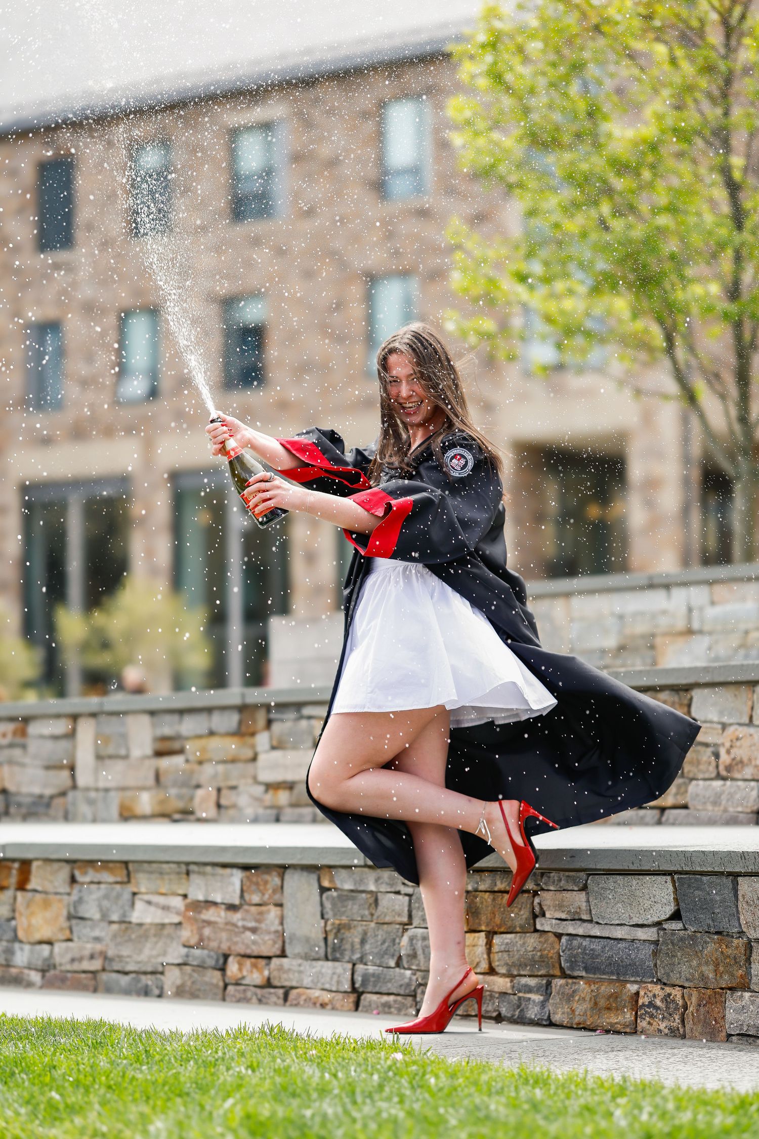 Person in black dress and red heels poses playfully on stone wall near brick building.