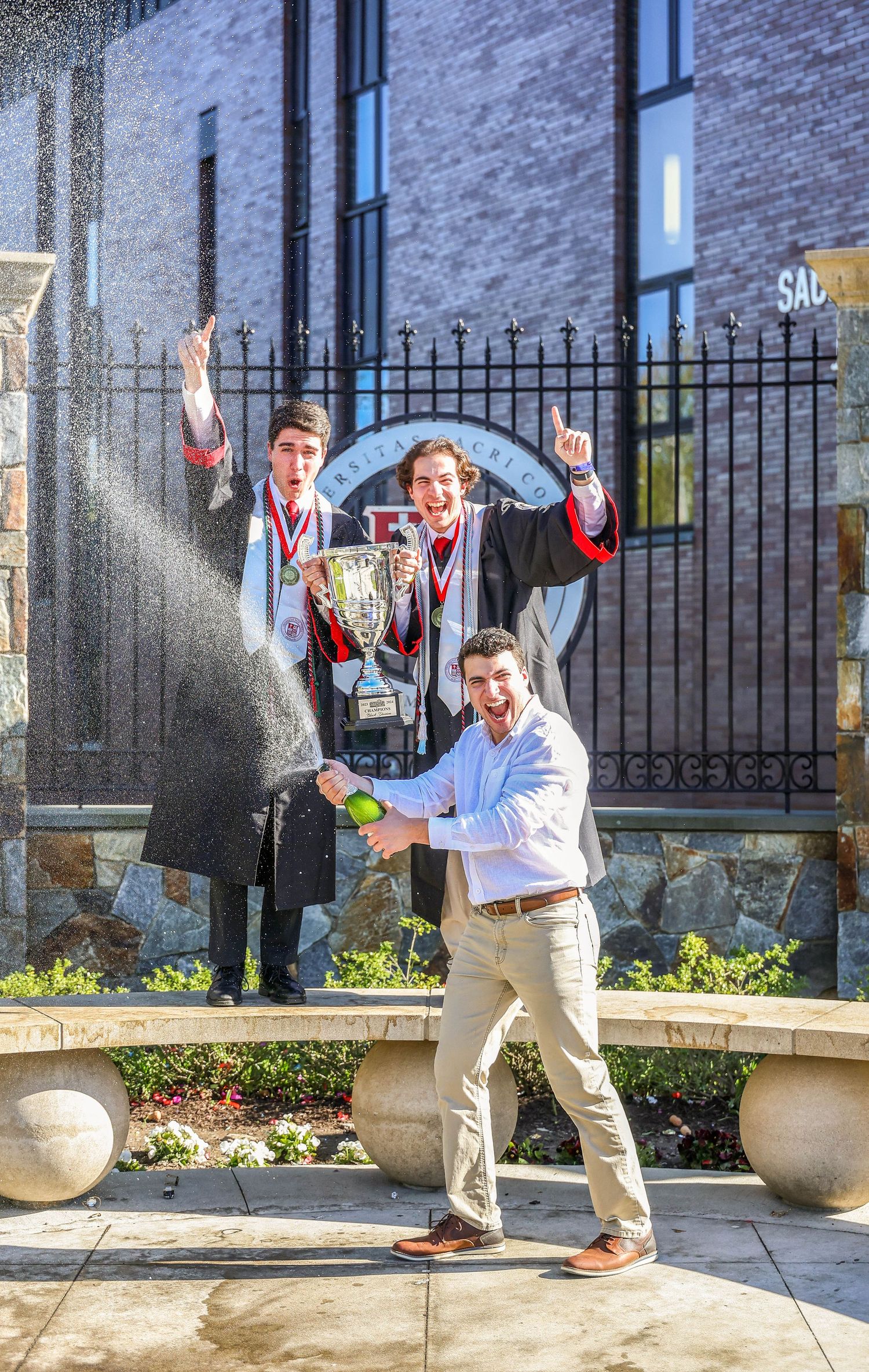 Graduates in formal attire leap and celebrate joyfully in front of brick building.