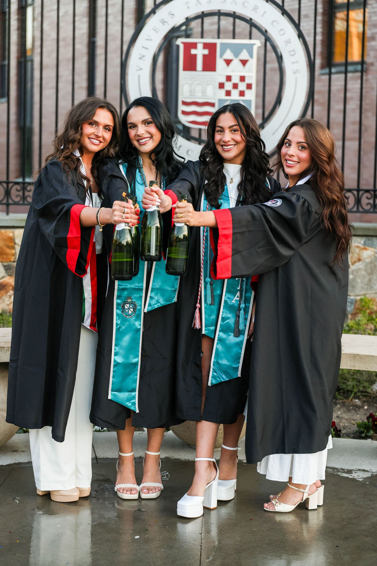 Graduates in black gowns with teal sashes show off their shoes in a fun group photo lineup.