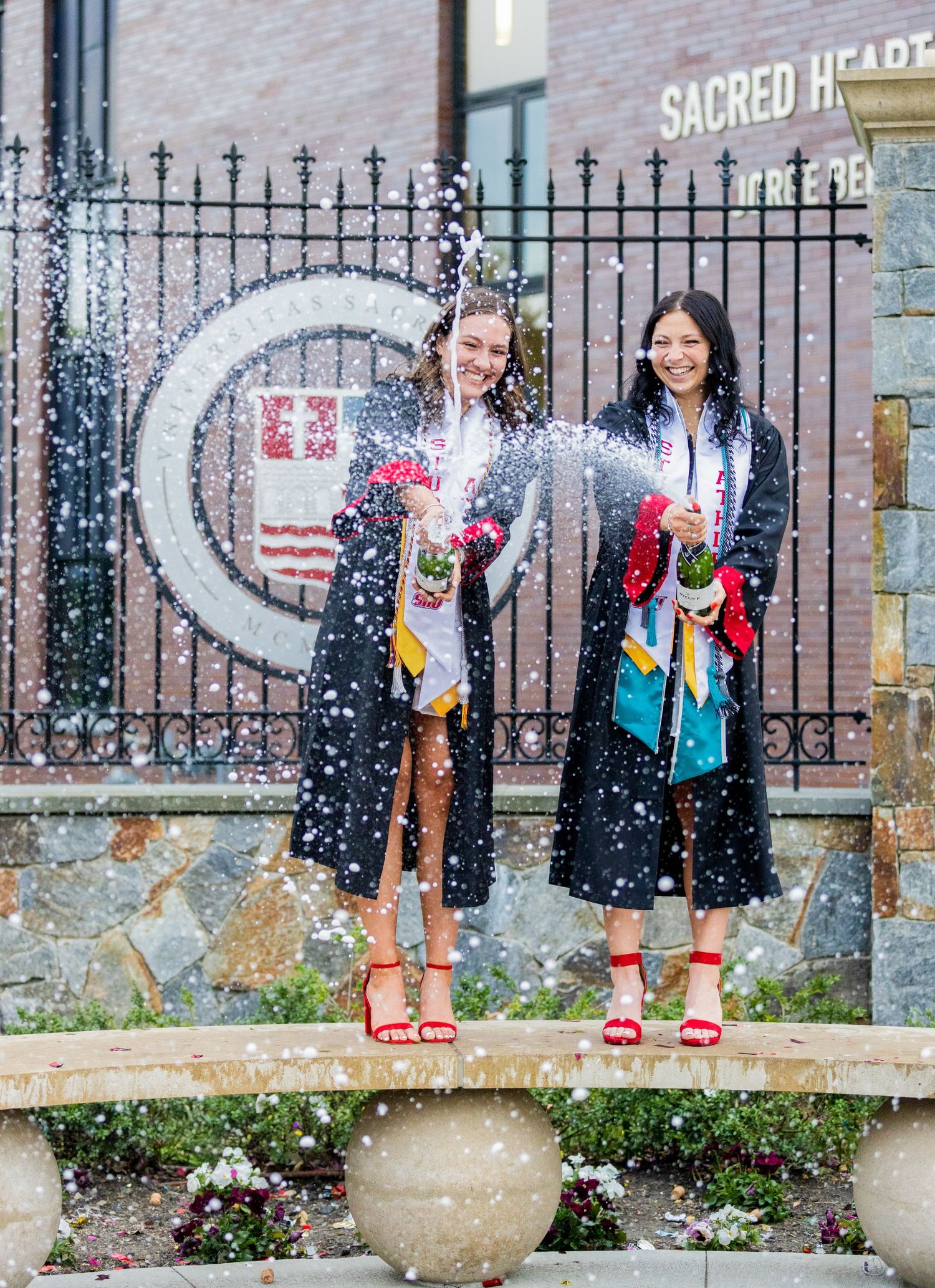 Graduates in traditional black gowns pose along a campus railing with sparklers creating a festive light effect.